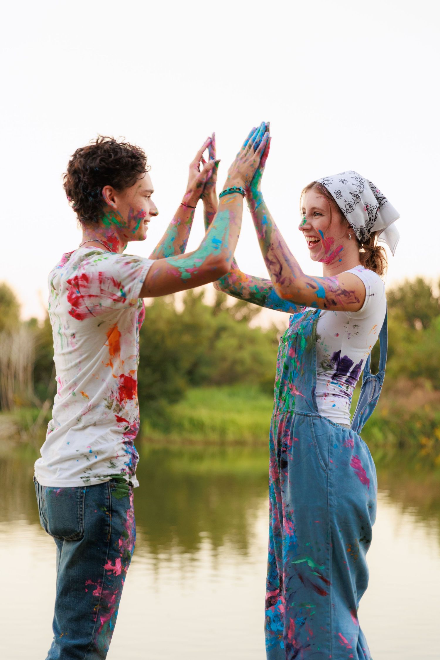 Paint-covered couple high-fiving and celebrating by a lake during a colorful outdoor photoshoot.
