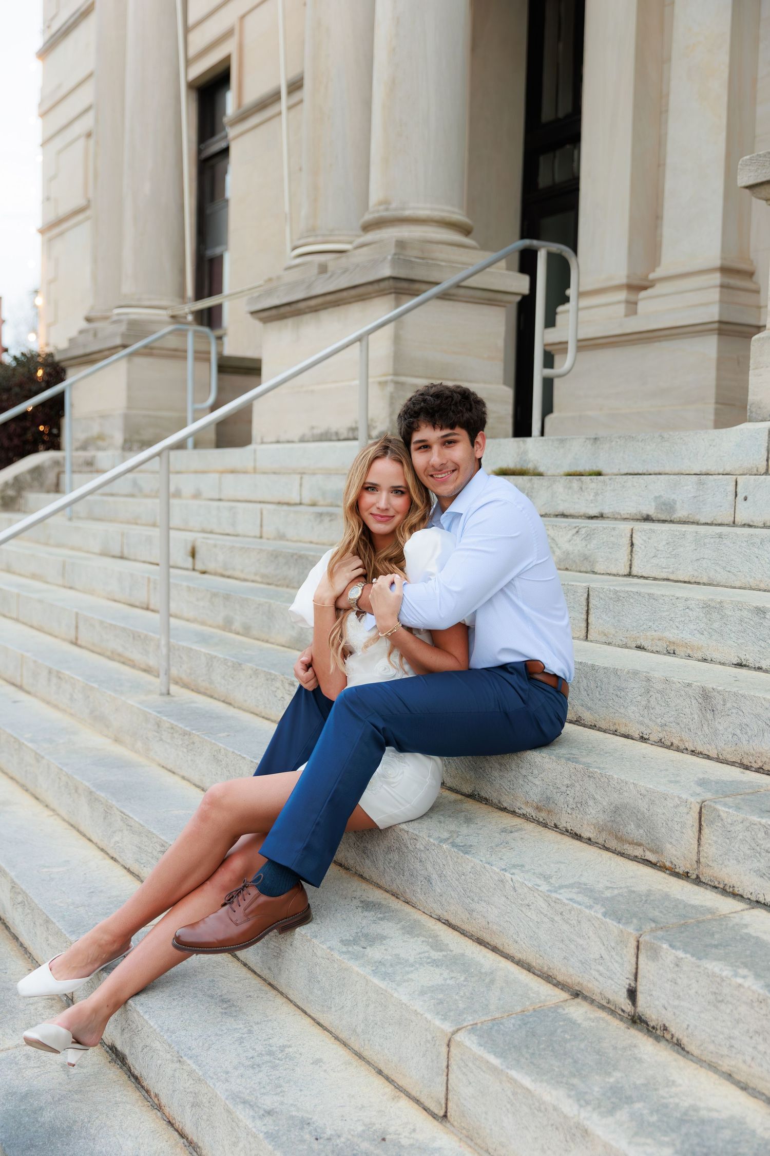 A couple sits playfully on stone steps of a grand building wearing casual summer attire.