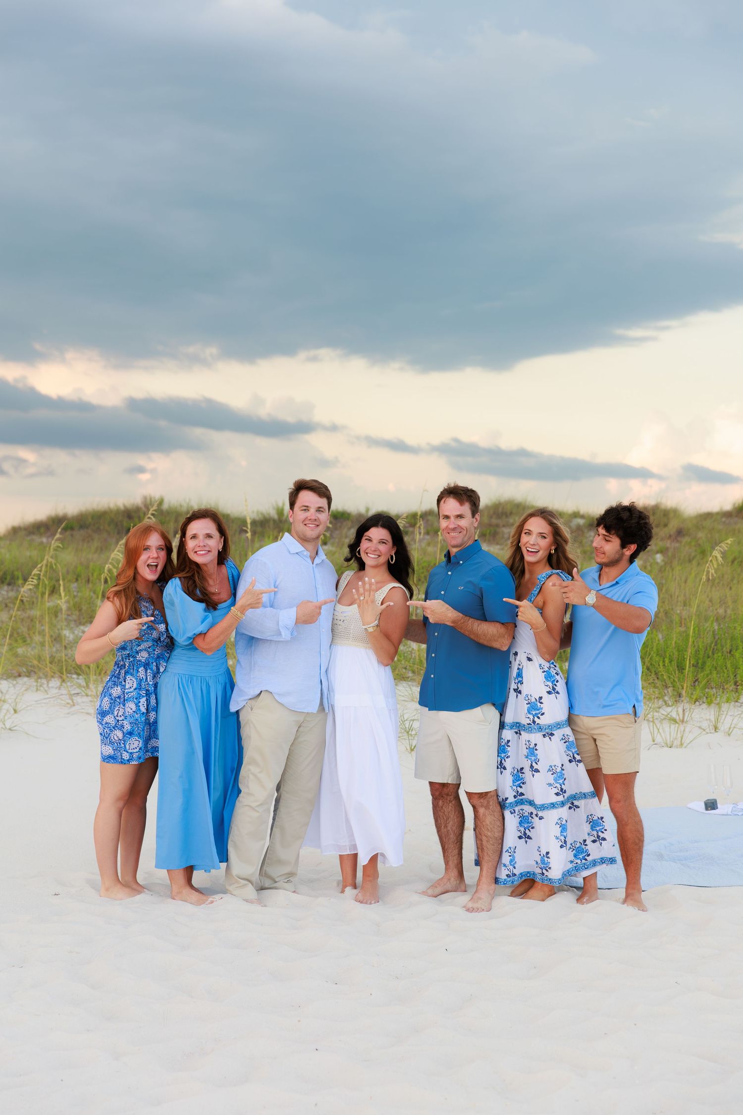 A group wearing coordinated blue and white outfits poses playfully on a white sandy beach with dunes in the background.