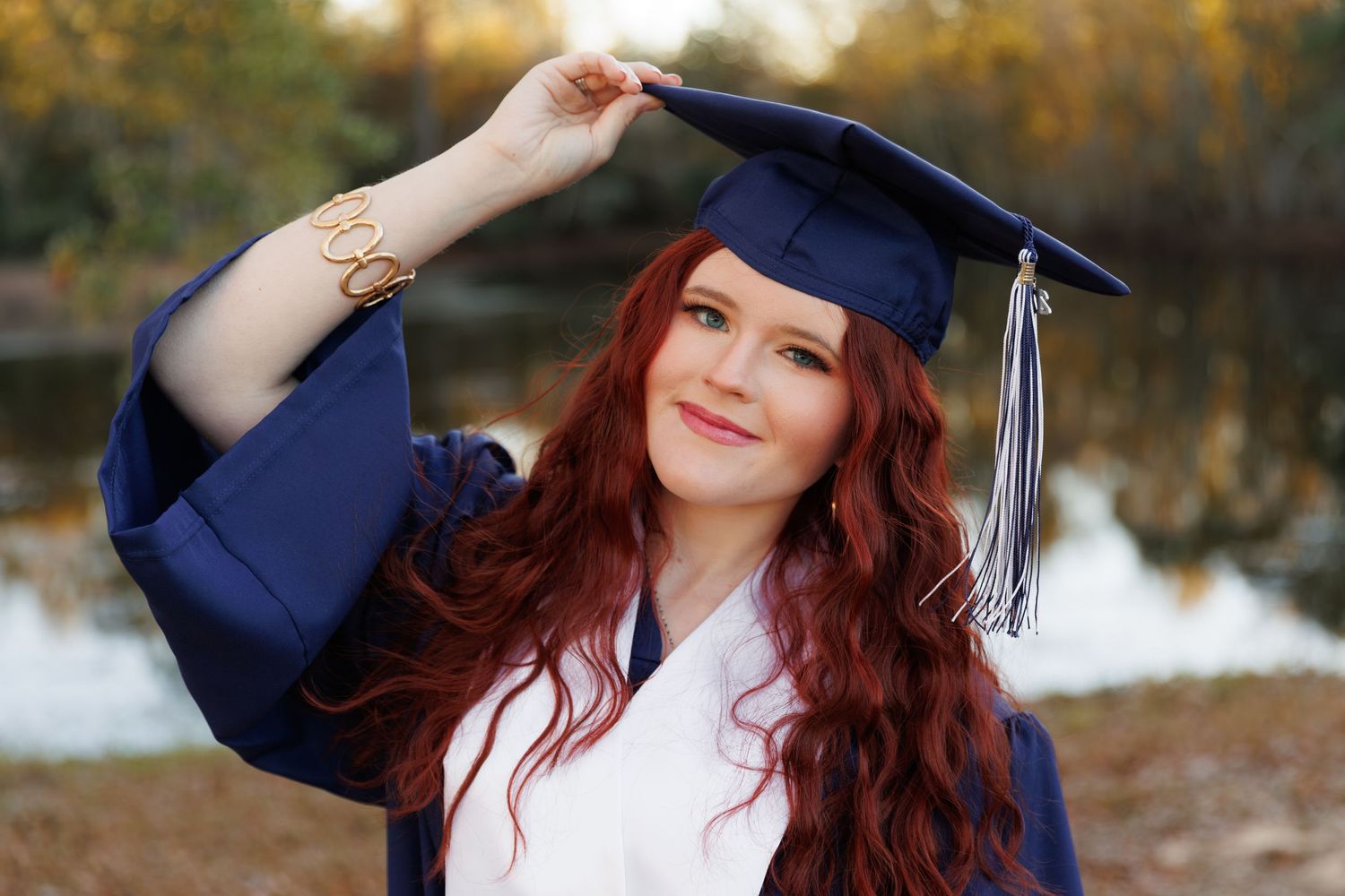 Graduate in navy blue cap and gown poses outdoors during fall season with natural background.