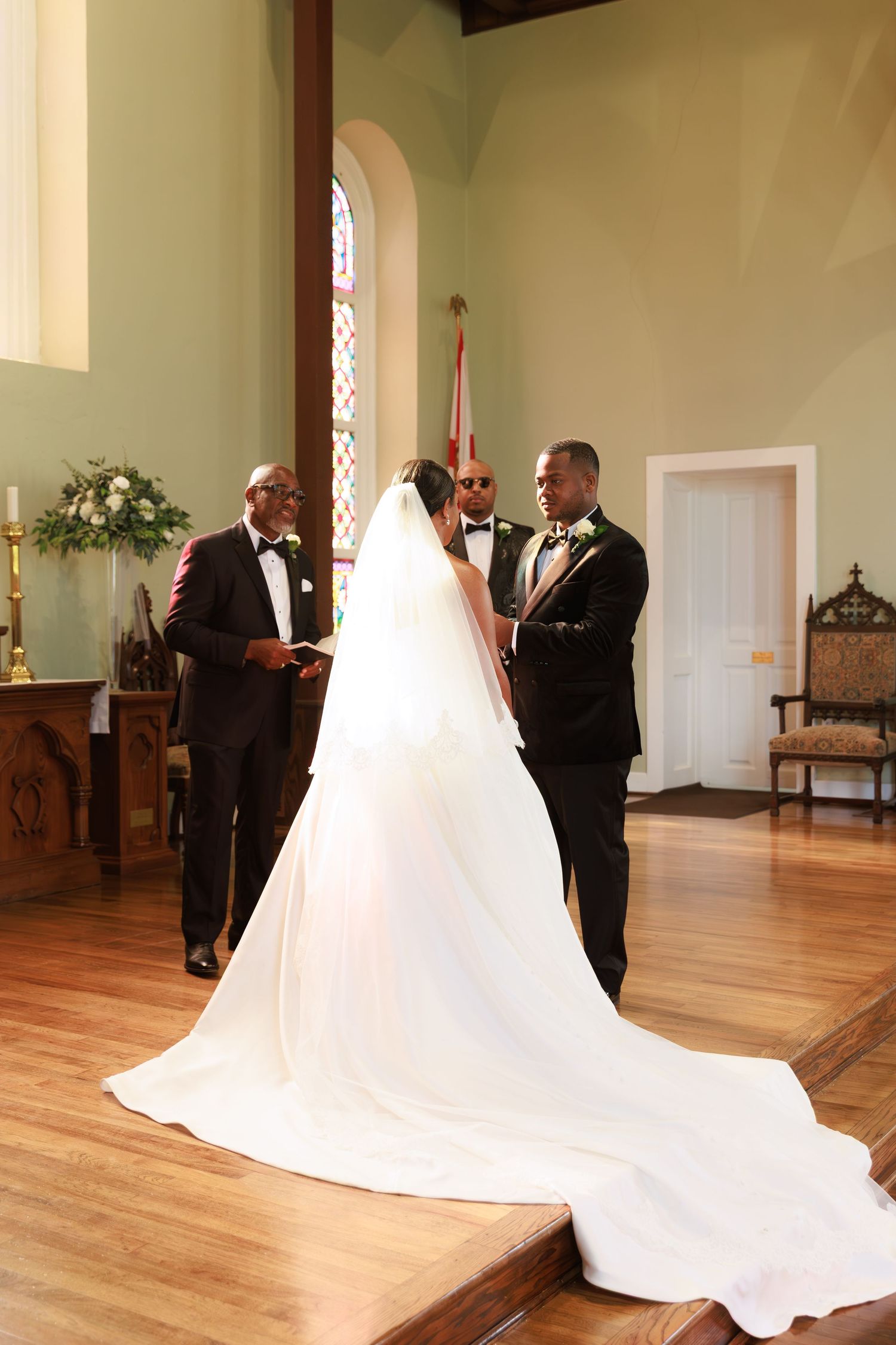 A wedding ceremony takes place in a bright church sanctuary with hardwood floors and a flowing white bridal gown.