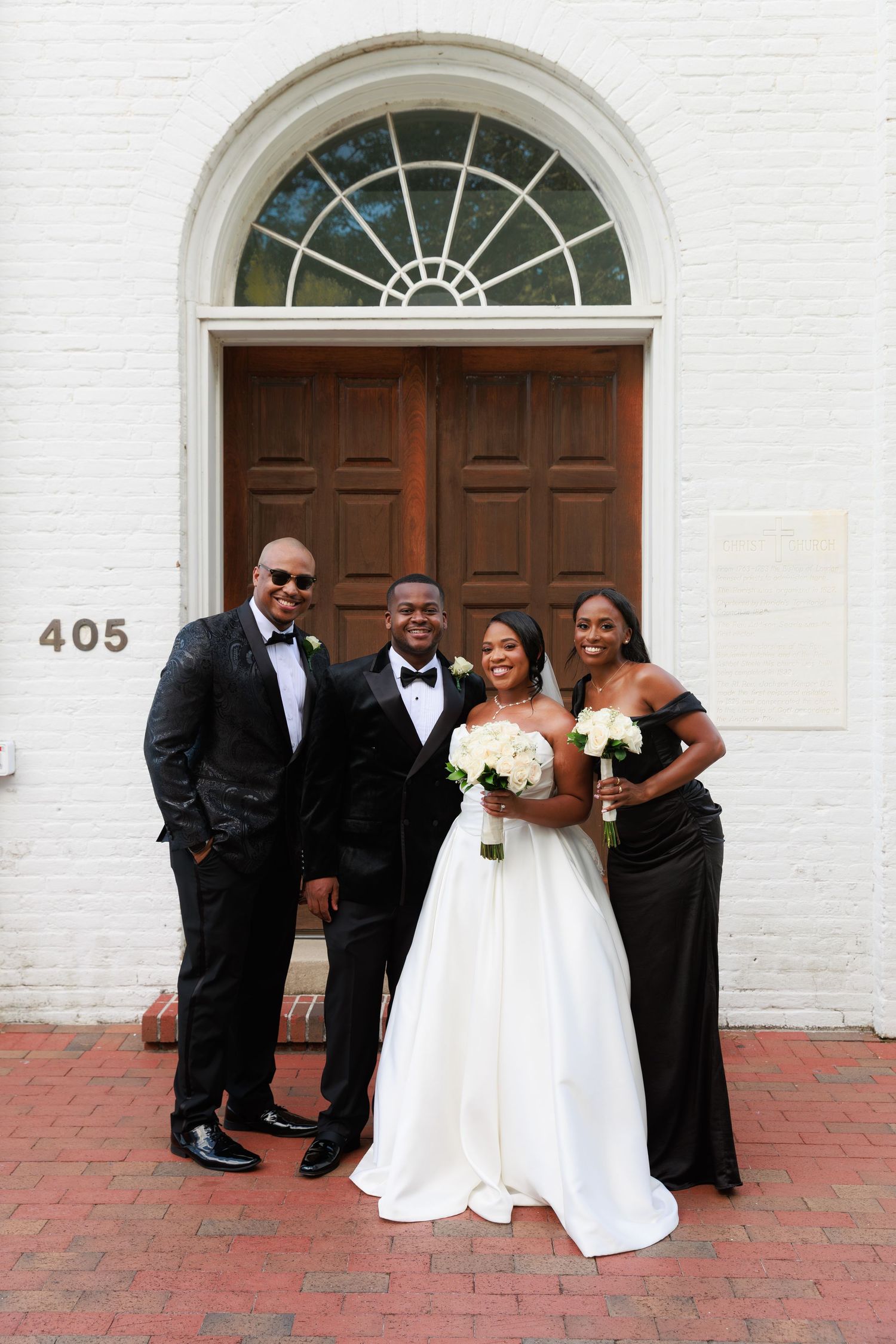 Wedding party in formal black and white attire poses together on brick patio outside historic white building.