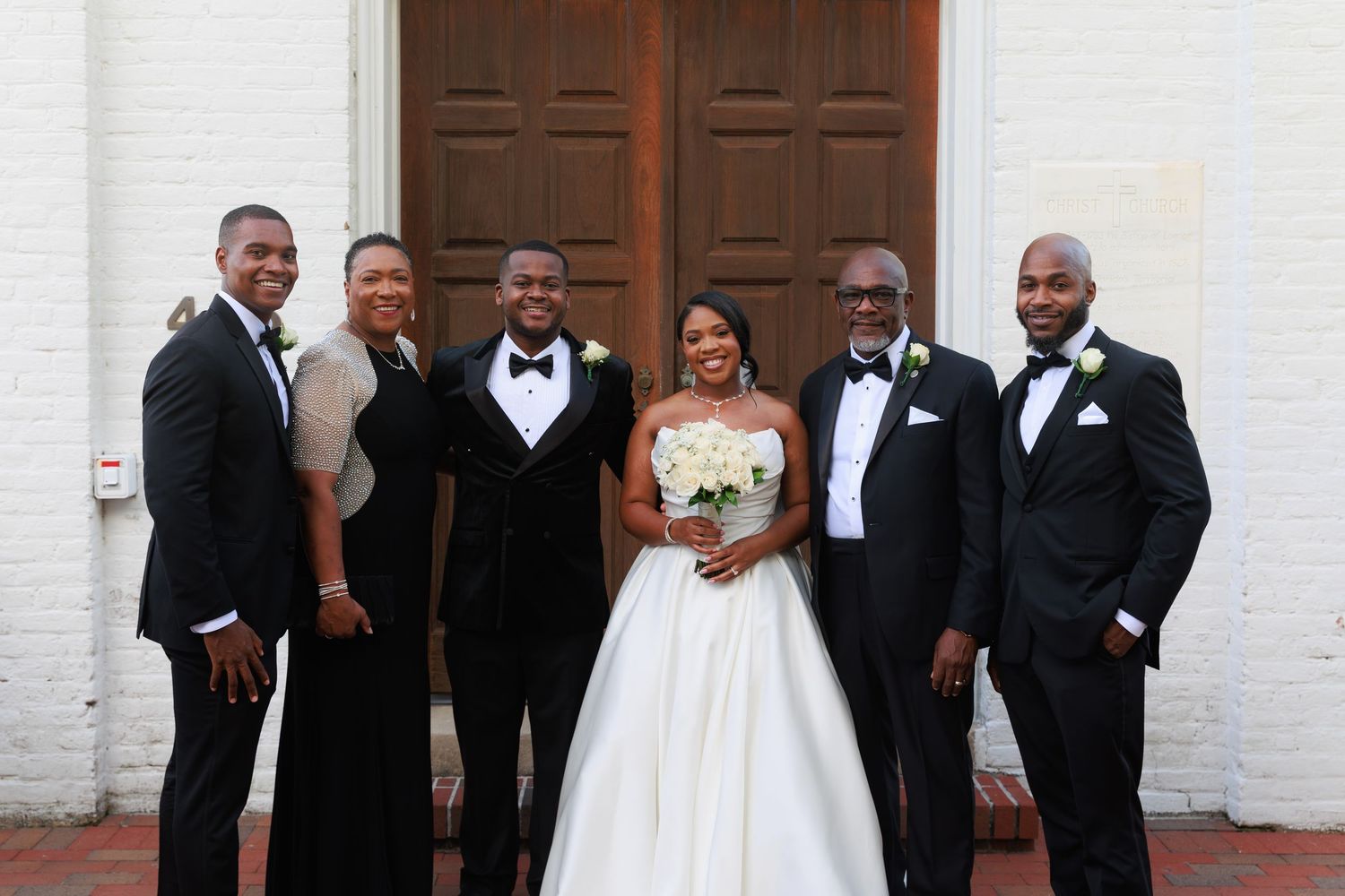 Wedding party members in black tuxedos and white gowns pose together on brick steps outside a white building.