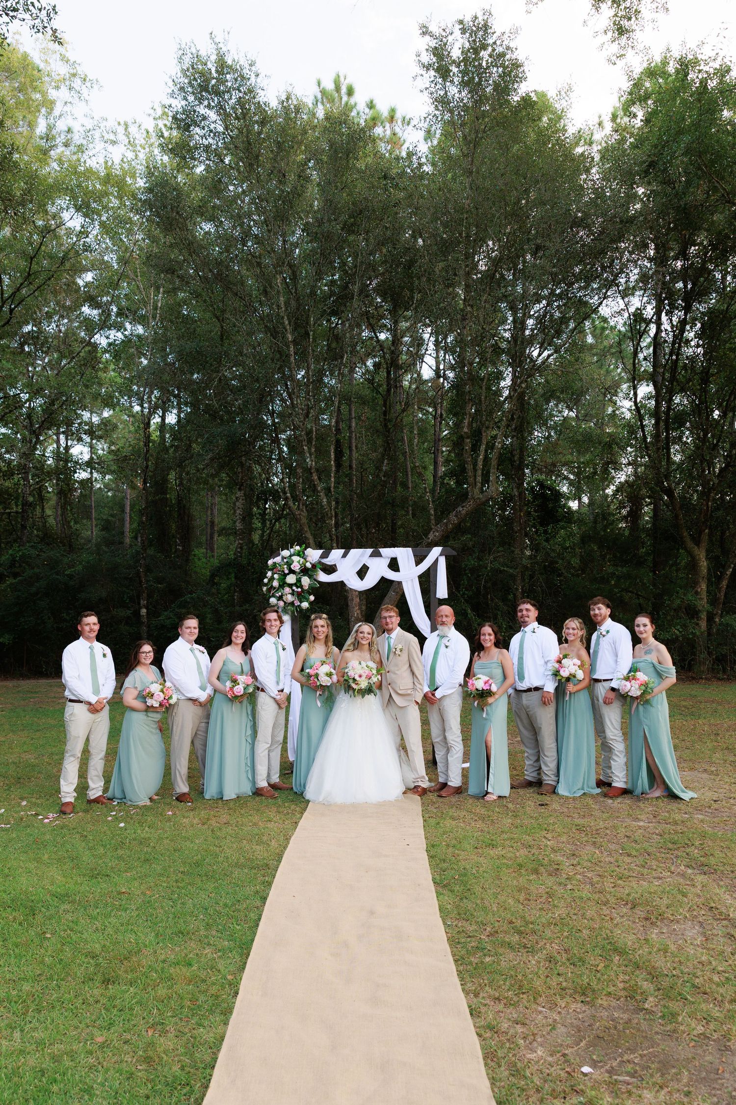 Wedding party in light blue dresses and khaki suits poses in line formation on outdoor path with trees in background.