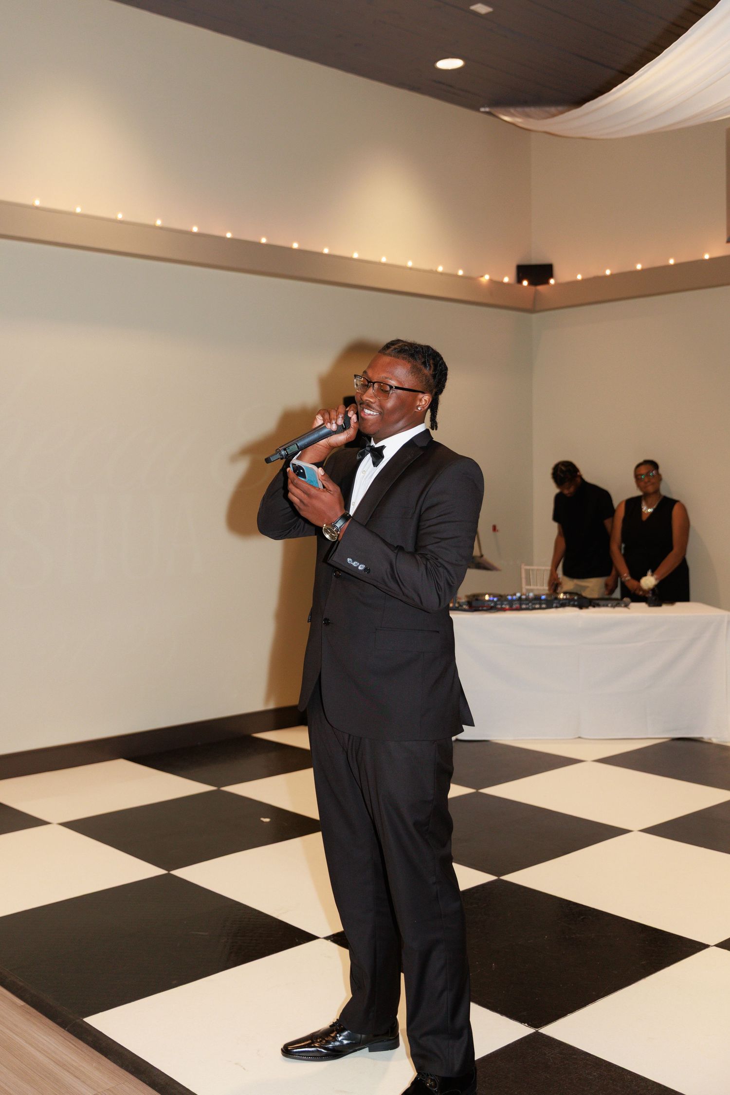 A person in a black tuxedo gives a speech while standing on a black and white checkered dance floor.