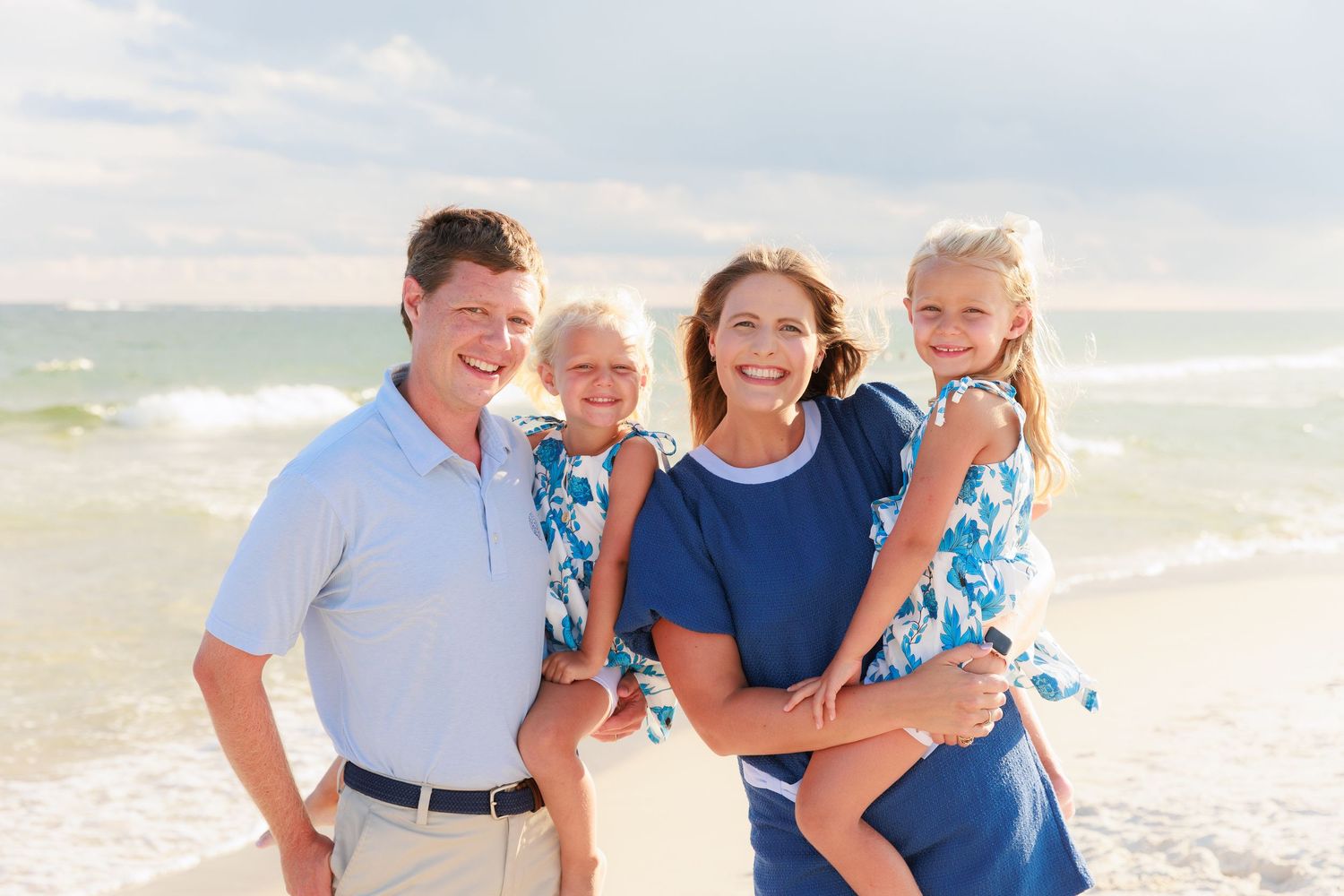 Happy family of four posing on a beach, parents holding two young girls in matching blue floral outfits.