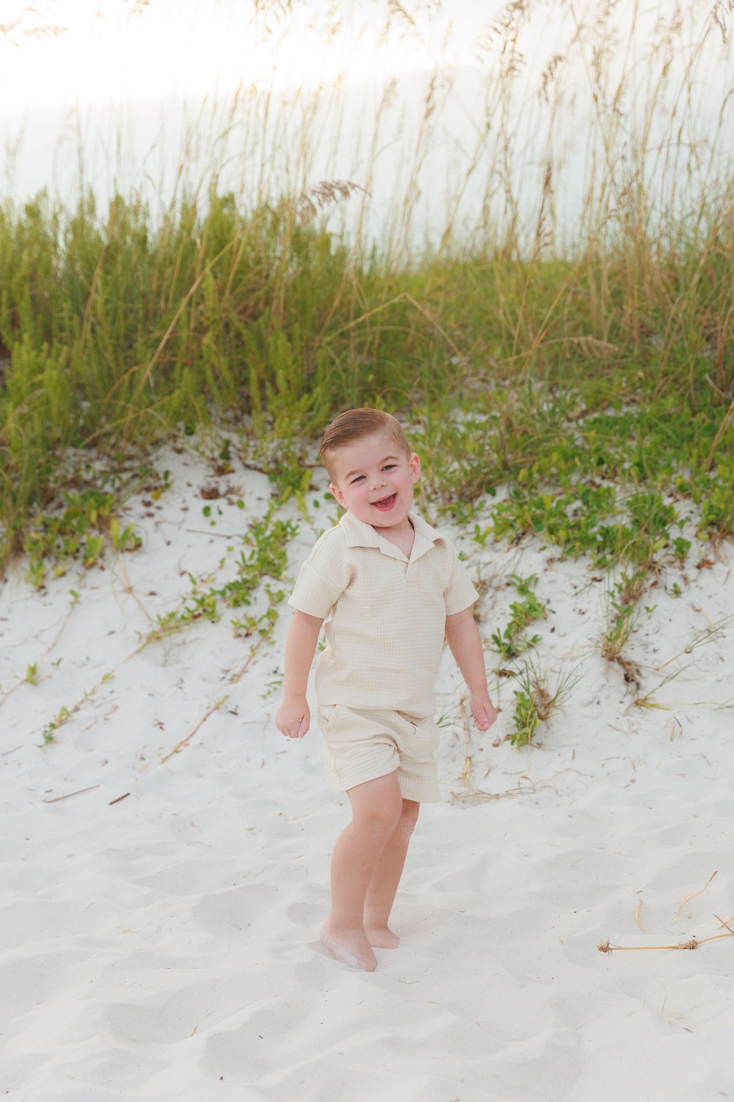 A joyful toddler boy in beige shorts set laughing on white sand beach with sea oats in background.