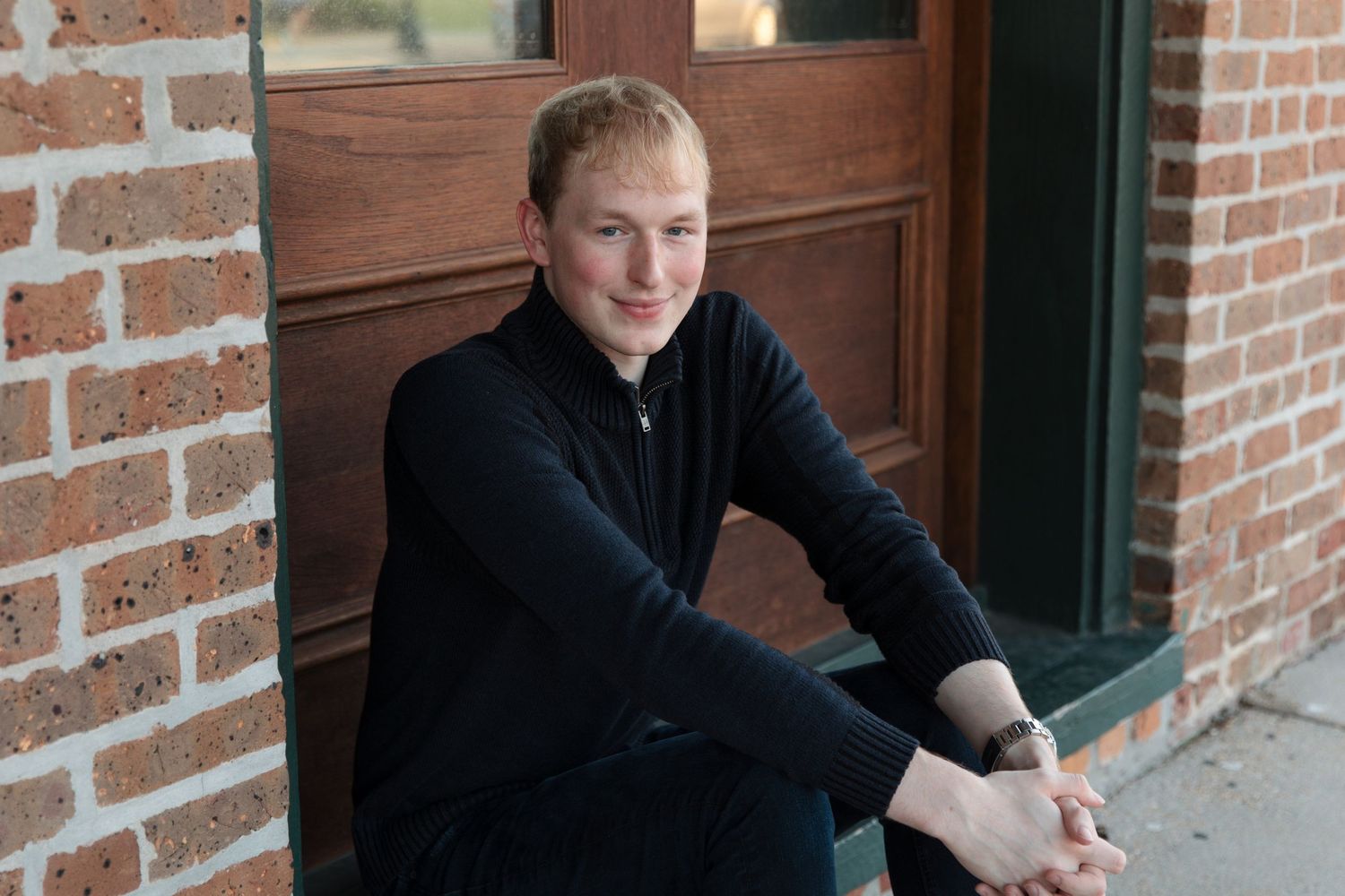 Young man in black jacket sitting on steps outside a brick building with a wooden door.