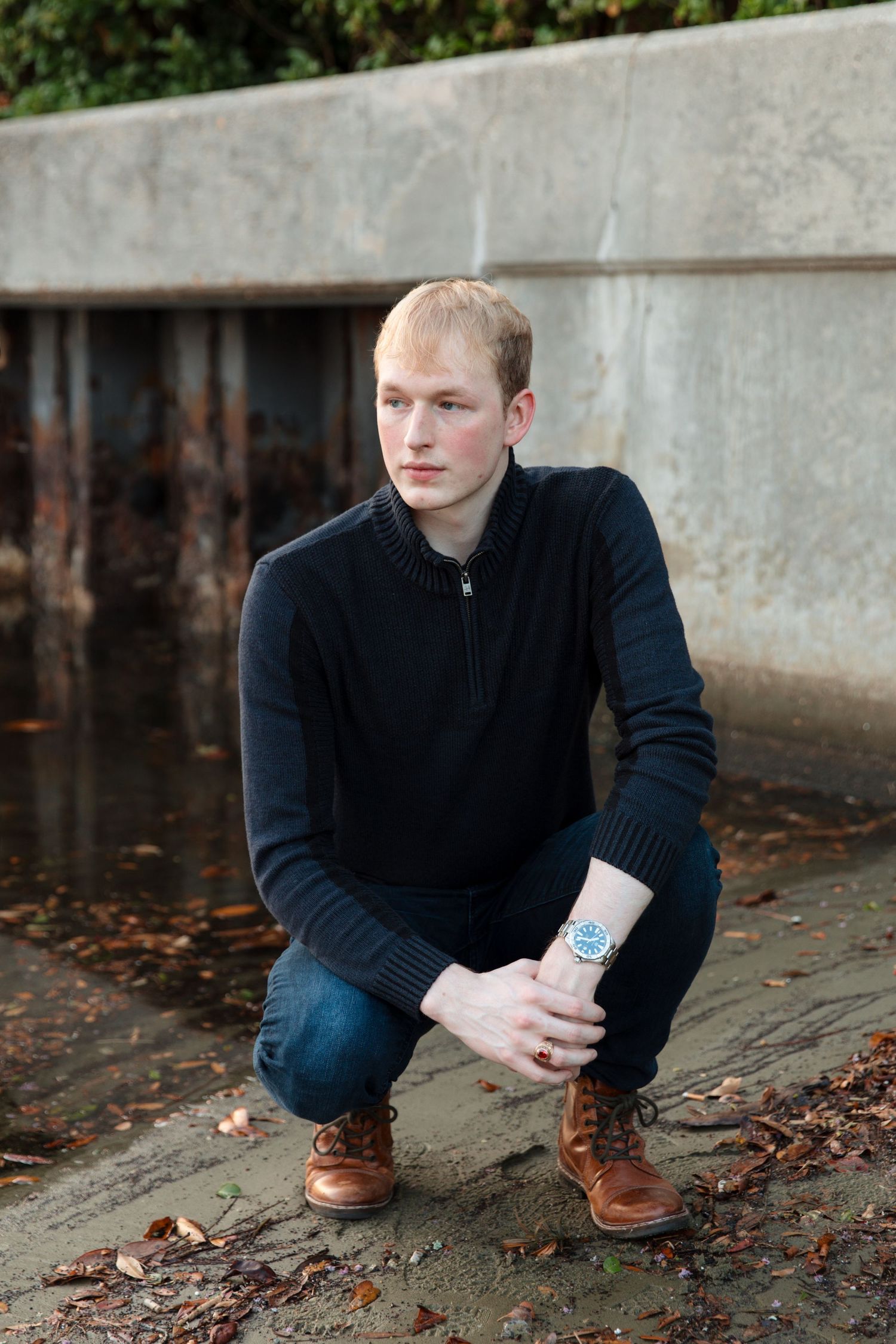 Young man in black sweater and jeans crouching outdoors near concrete wall wearing brown boots and blue watch.