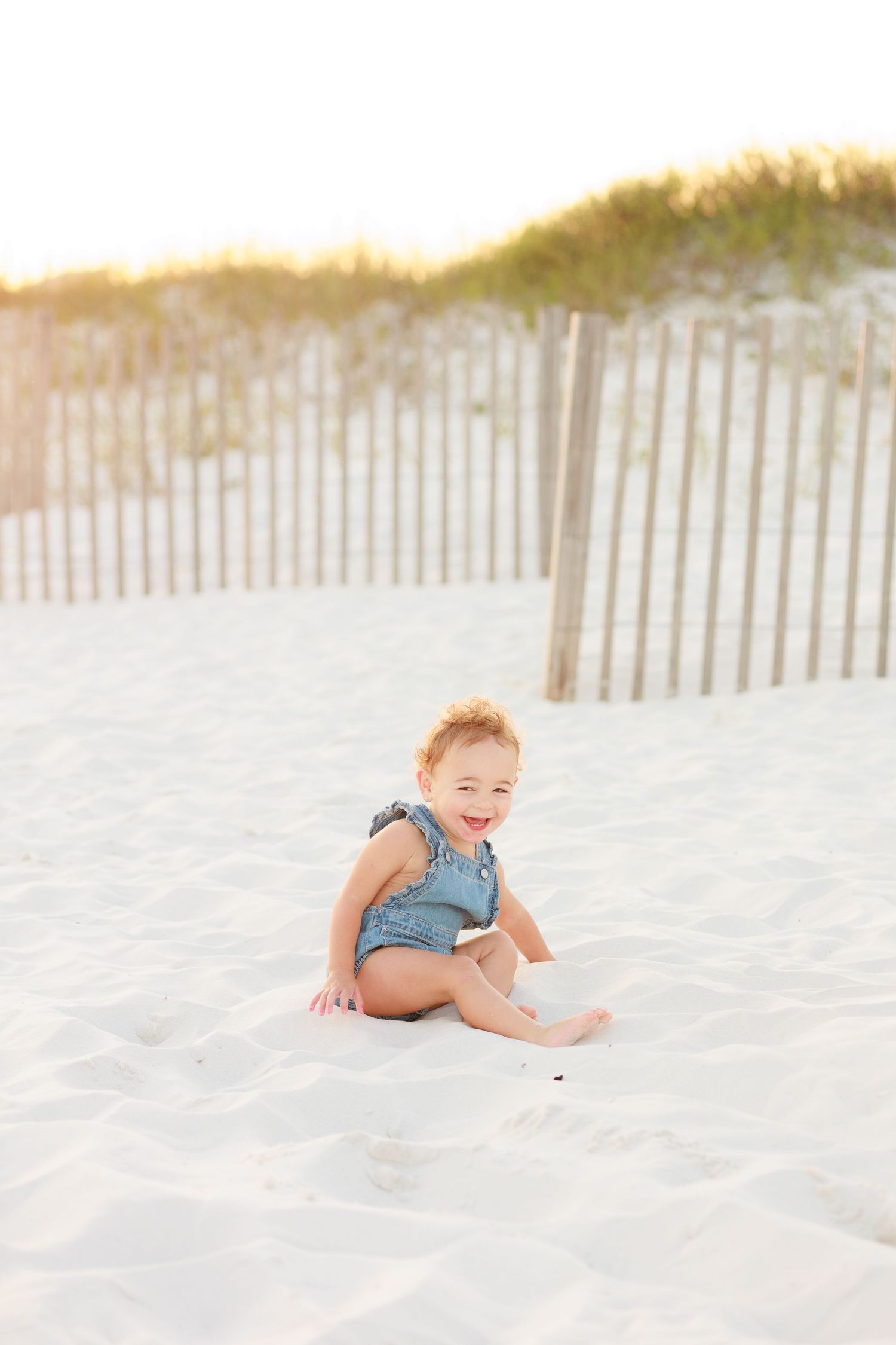 Happy toddler in denim overalls sitting and smiling on white sand beach near wooden fence.