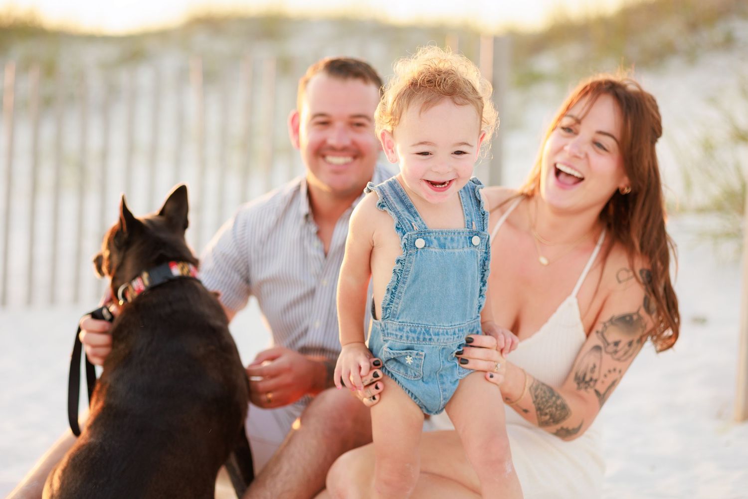 Happy family with a toddler in denim overalls and a black dog smiling on a sandy beach.