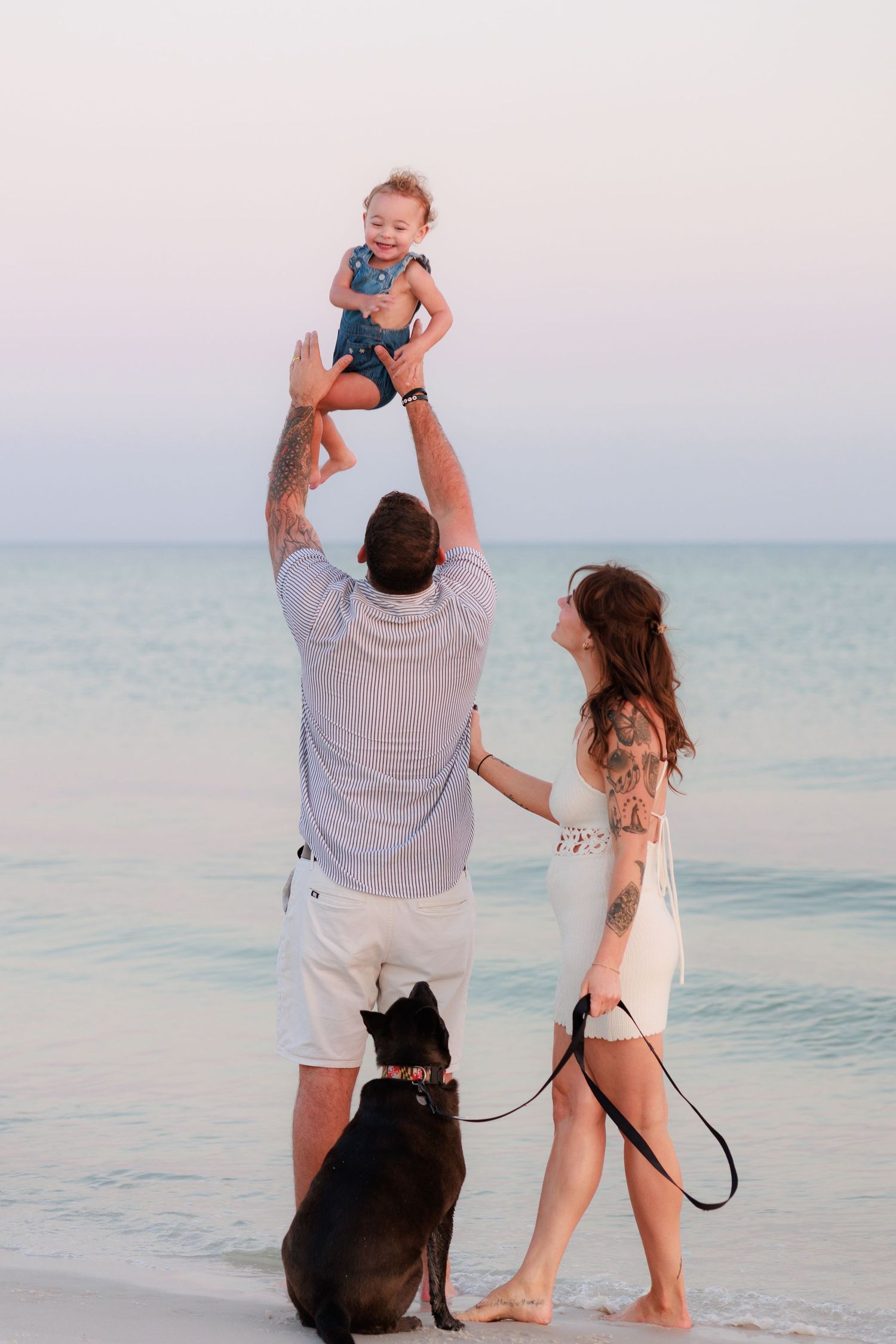 Family with tattoos tossing baby in air on beach at sunset with black dog on leash beside calm ocean.