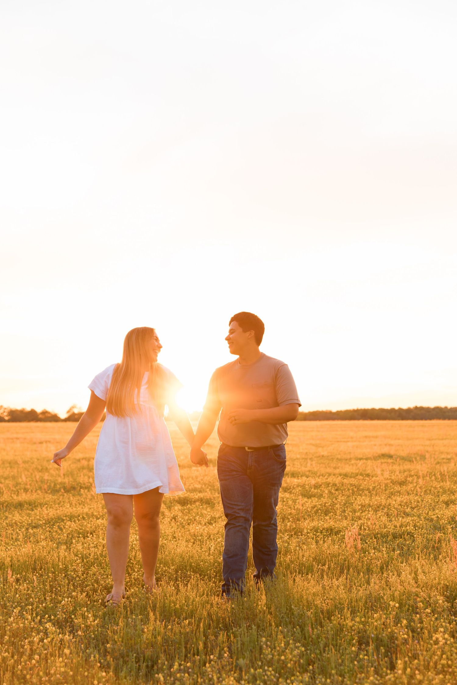 Couple holding hands walking through golden field at sunset, bathed in warm orange light.