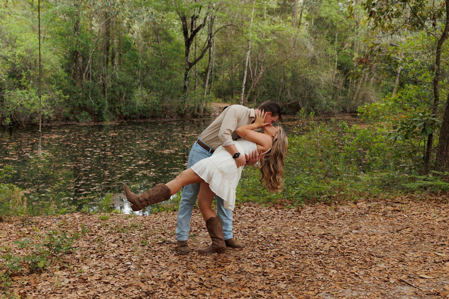 Couple sharing a romantic dip kiss near a tranquil forest pond surrounded by autumn leaves.
