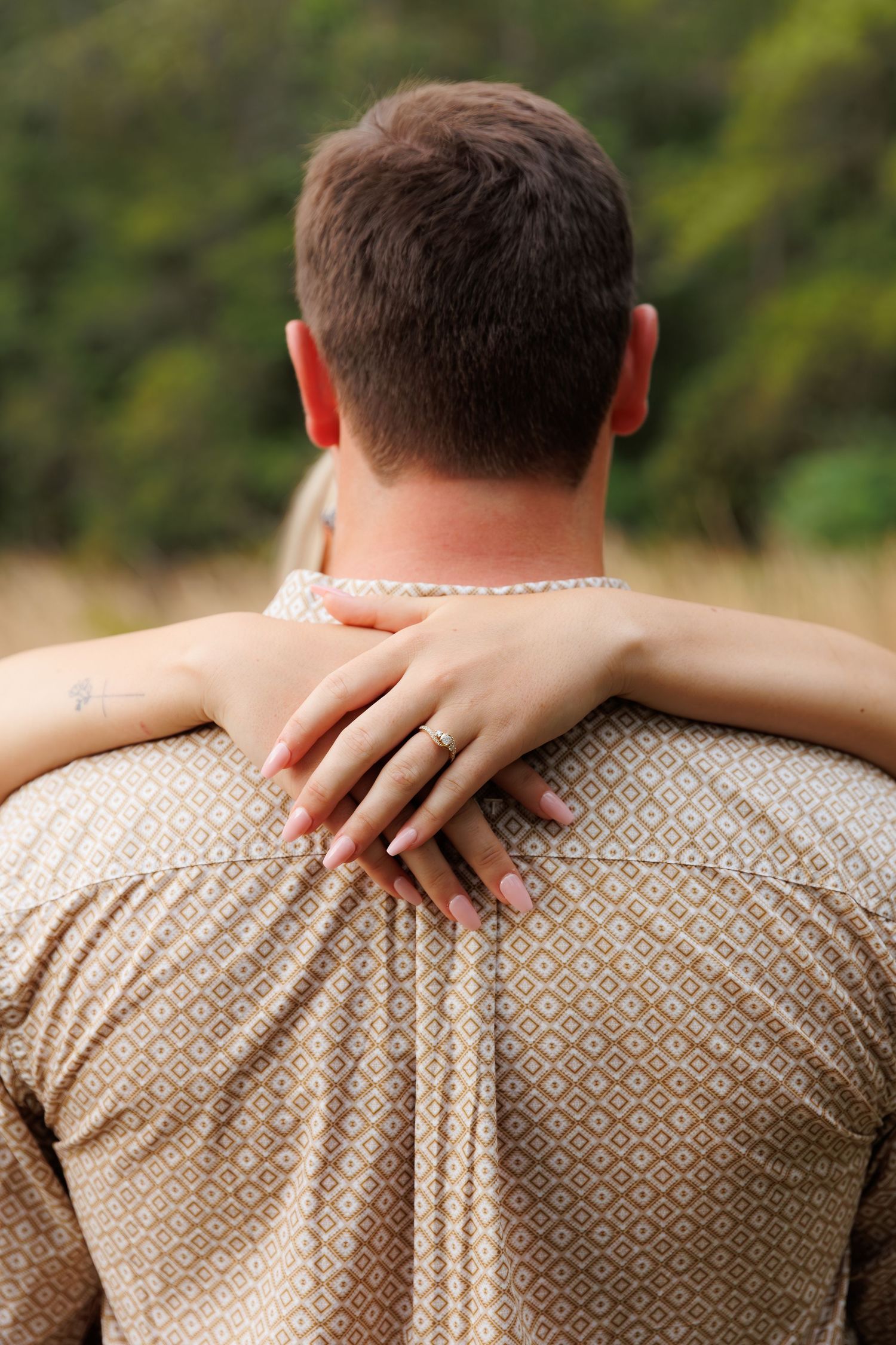 A woman with an engagement ring embraces a man from behind, her hands resting on his shoulders outdoors.