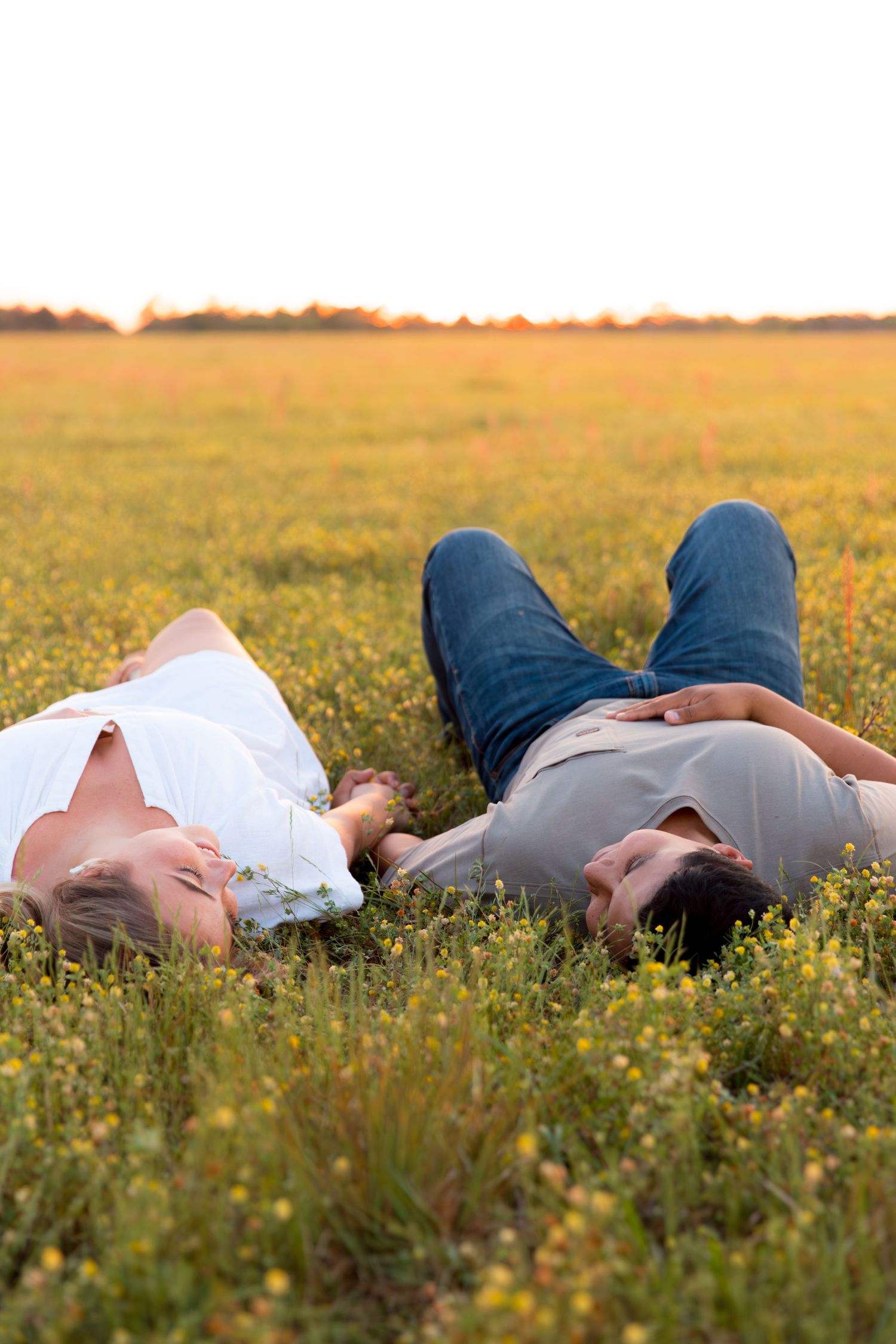 Couple lying in a sunny field of yellow wildflowers, smiling and holding hands at golden hour.