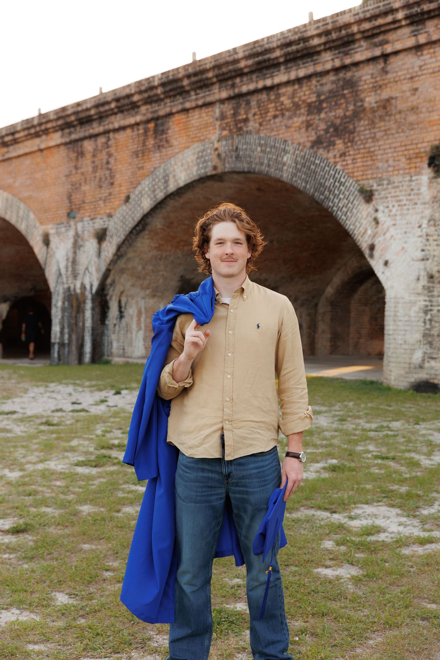 A smiling man with curly red hair holds a blue jacket over his shoulder, standing before a historic brick archway.