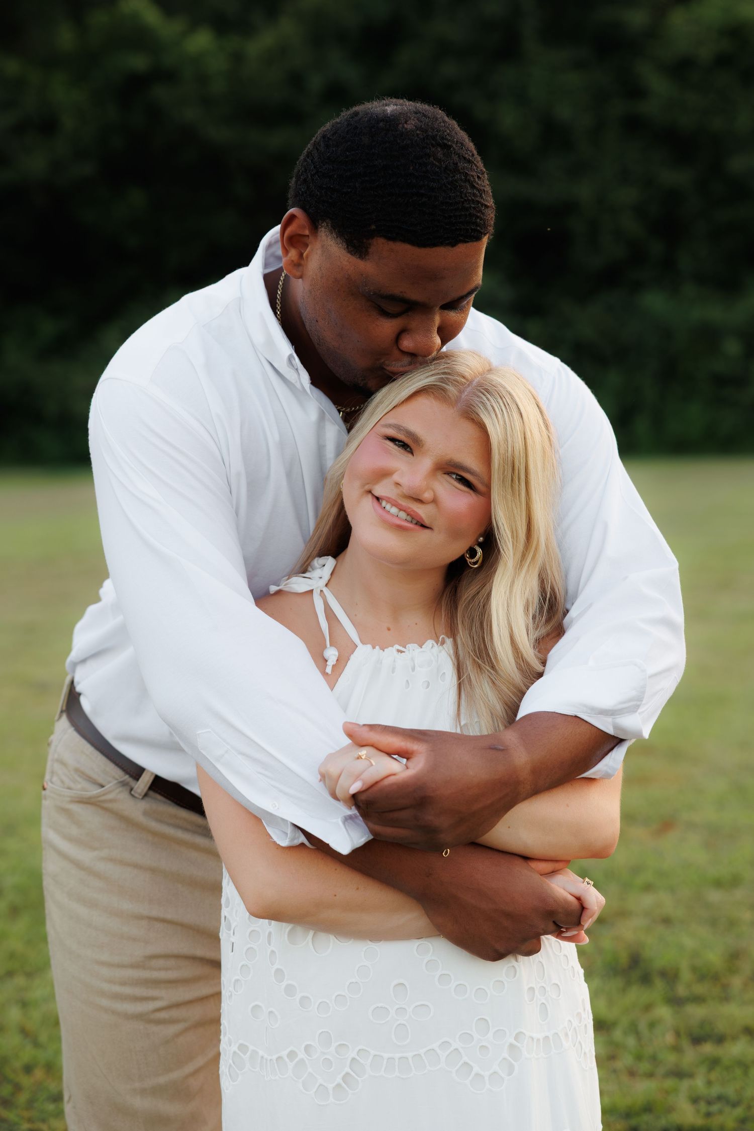 A man in a white shirt embraces a smiling blonde woman in a white dress outdoors, sharing a tender romantic moment.