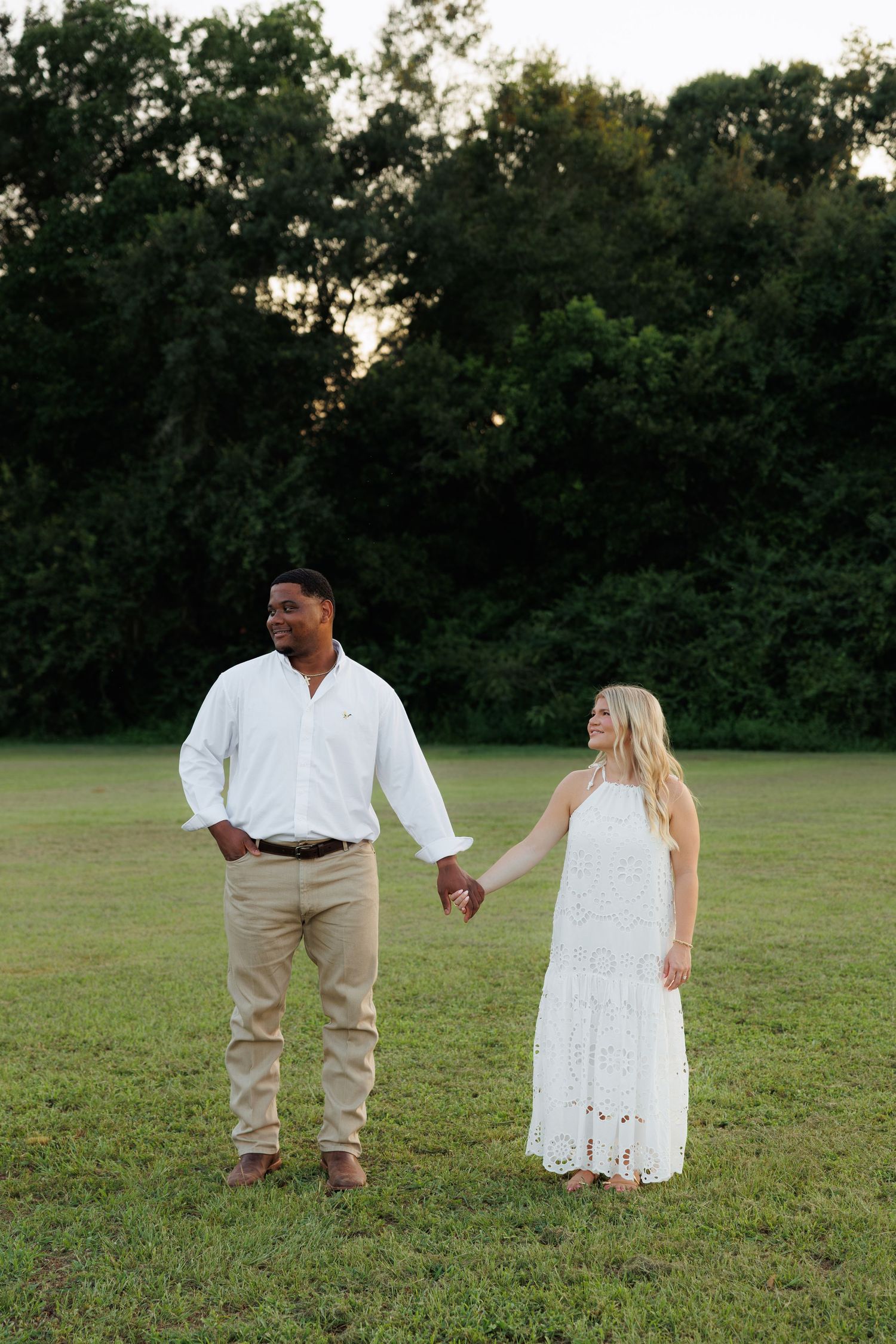 Couple holding hands in a lush green field at golden hour, man in white shirt and woman in lace dress.