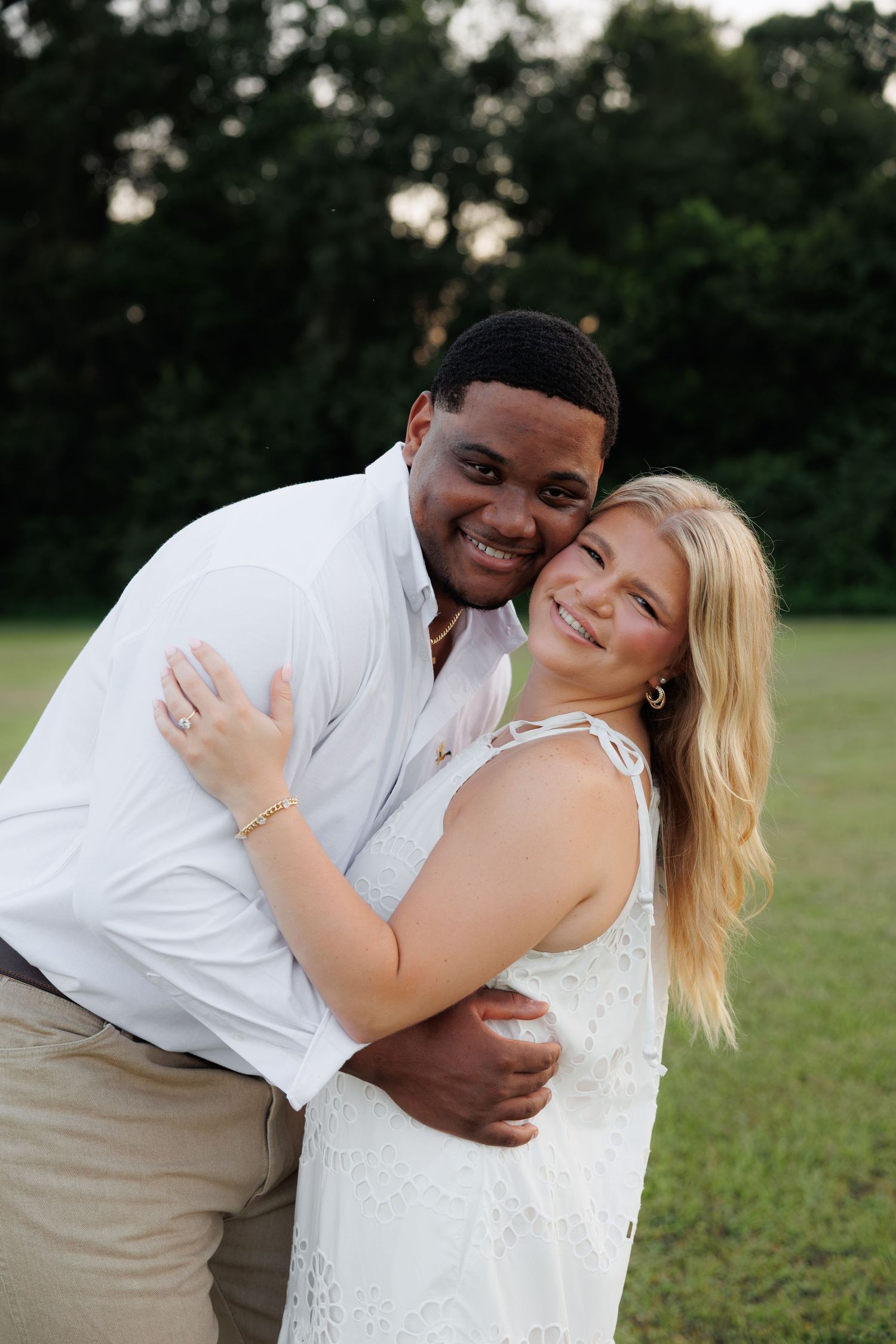 Smiling couple embracing closely in a green field at dusk, both dressed in white and khaki.