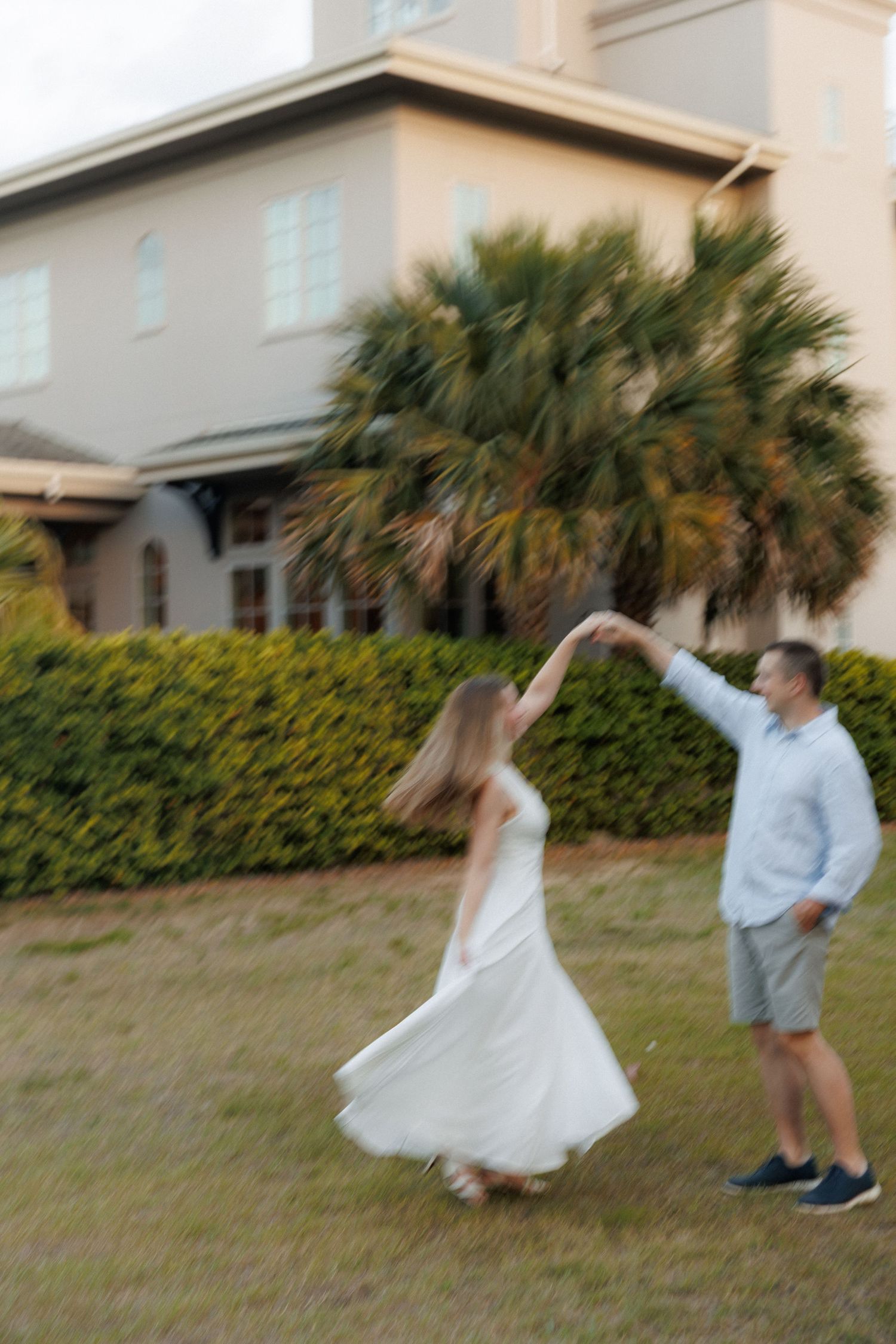 Motion-blur photo of a couple dancing outdoors, man spinning woman in a flowing white dress near palm trees.