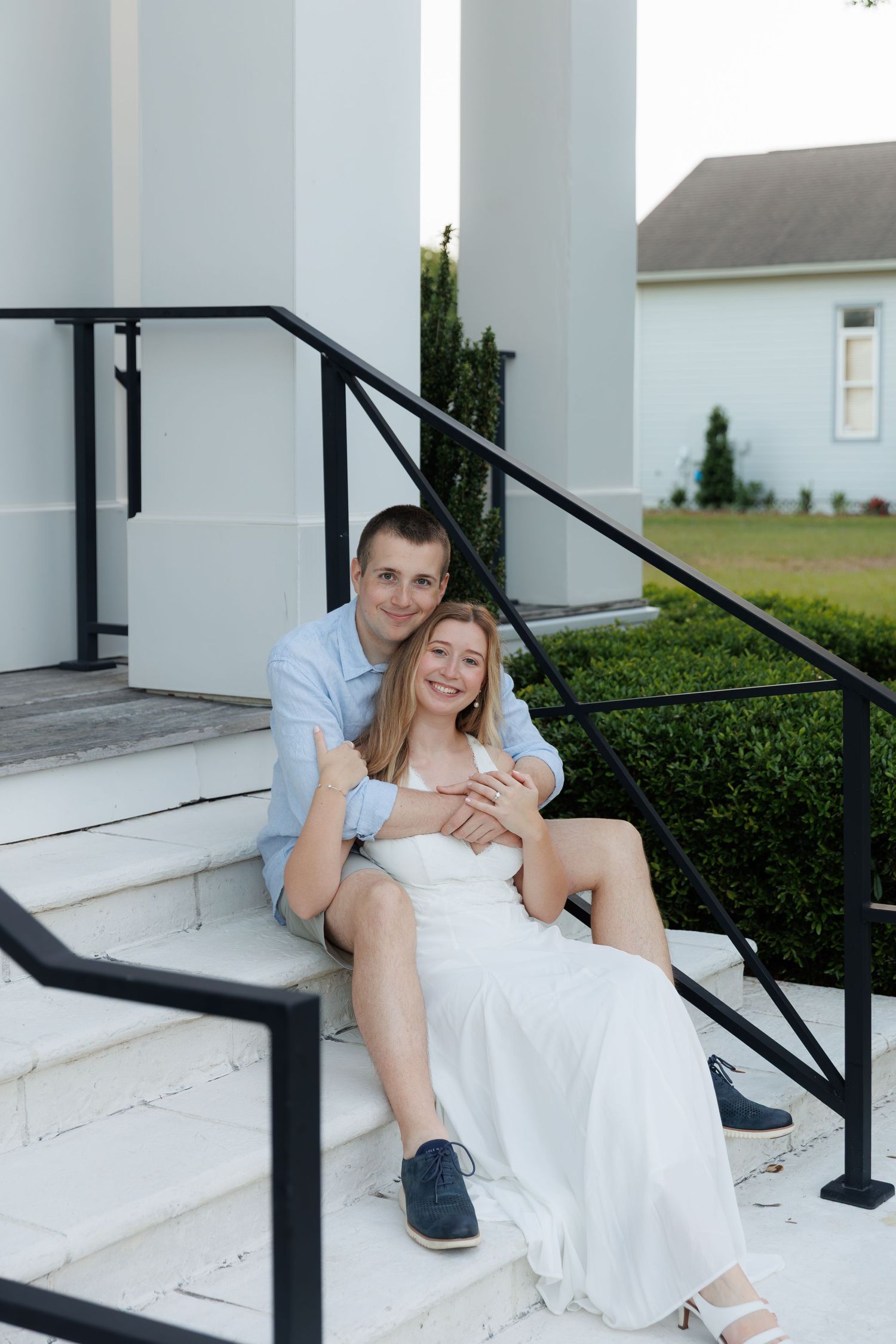 Smiling couple sitting closely on white building steps, man embracing woman in white dress from behind.