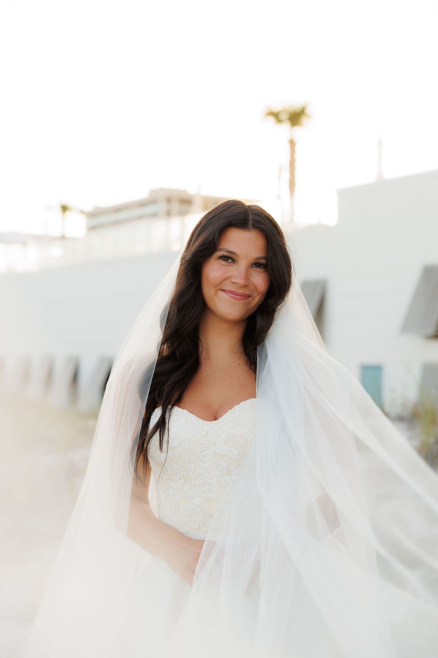 A smiling bride in a strapless white gown and flowing veil poses outdoors with palm trees in the background.
