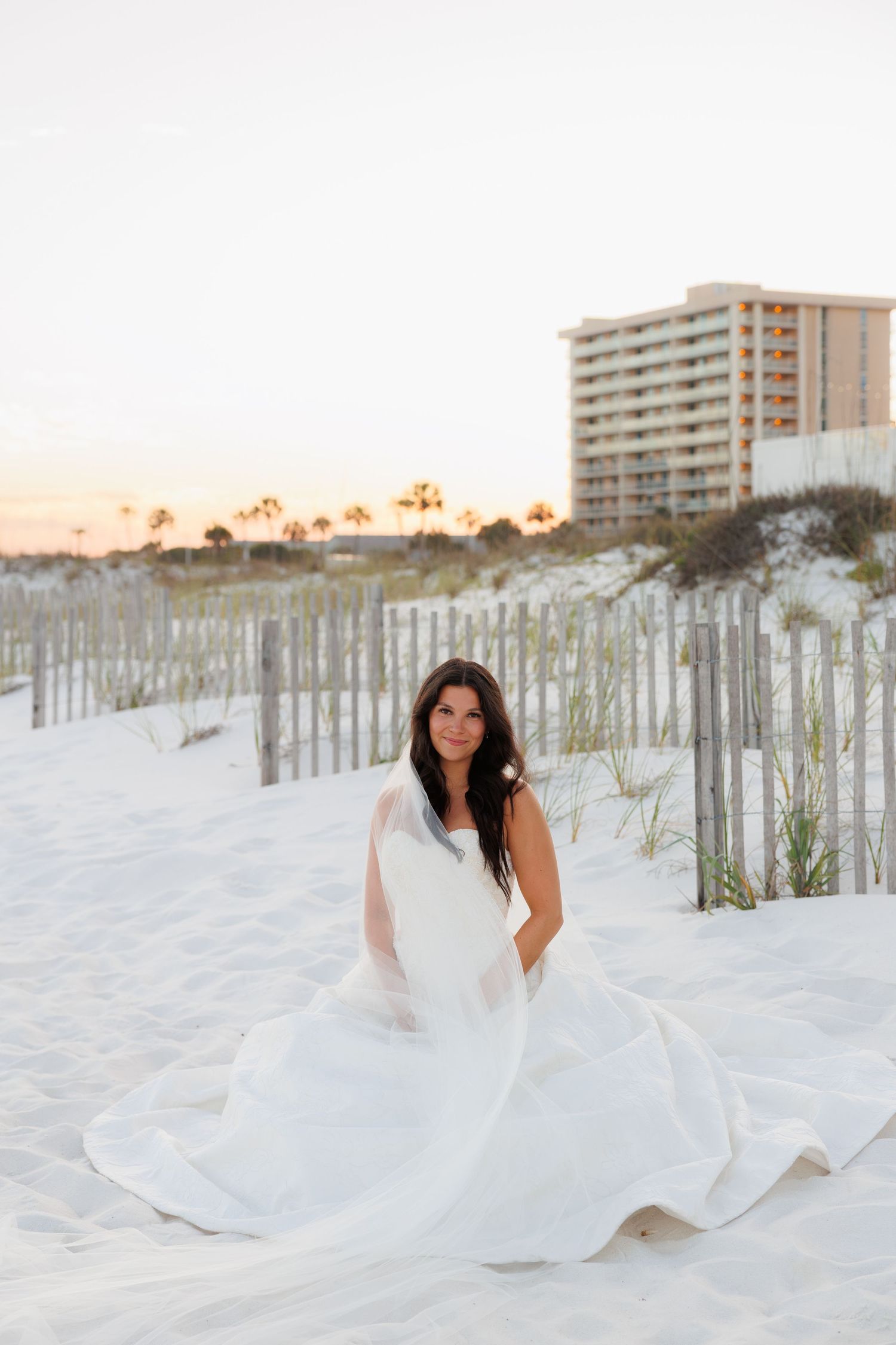 Bride in flowing white wedding dress sits on white sand beach at golden hour.