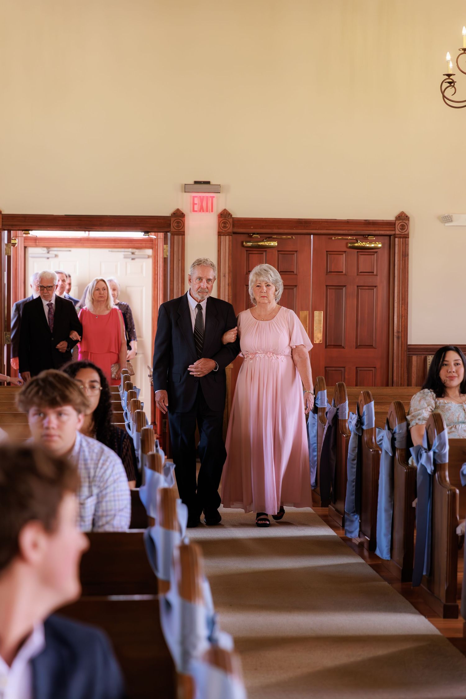 Processional in a church sanctuary with wood pews and doors, people walking down the center aisle during a ceremony.