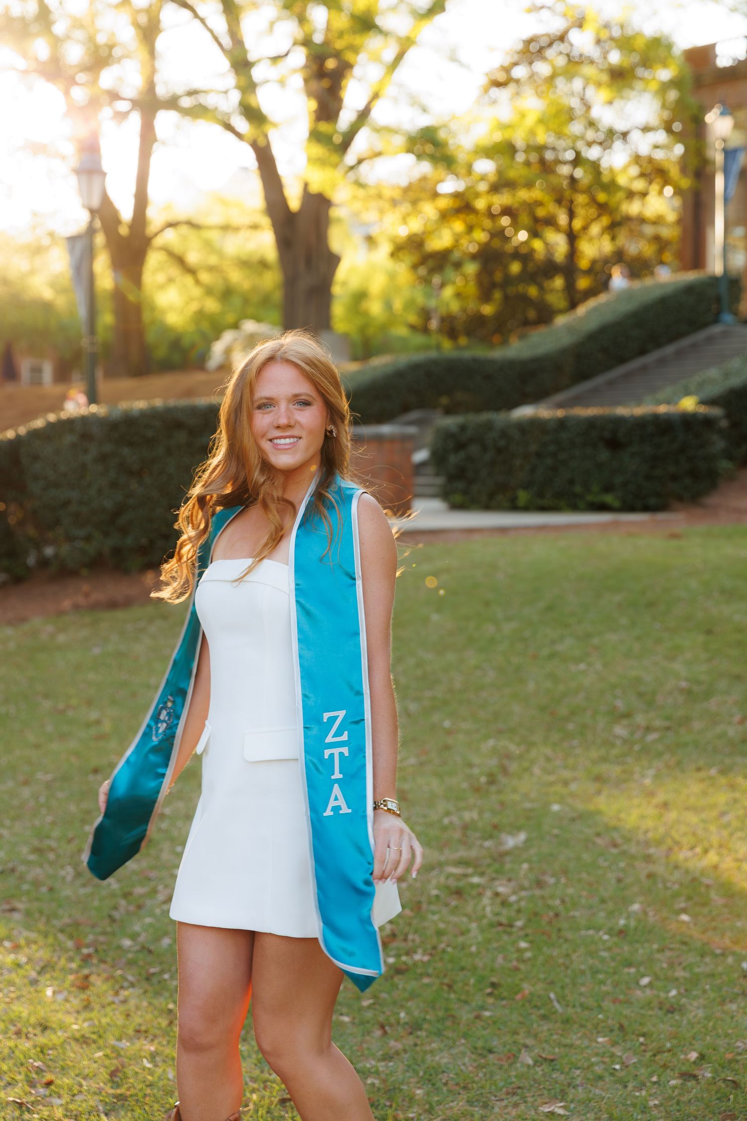 Smiling college graduate wearing ZTA sorority stole in white dress on campus at sunset.