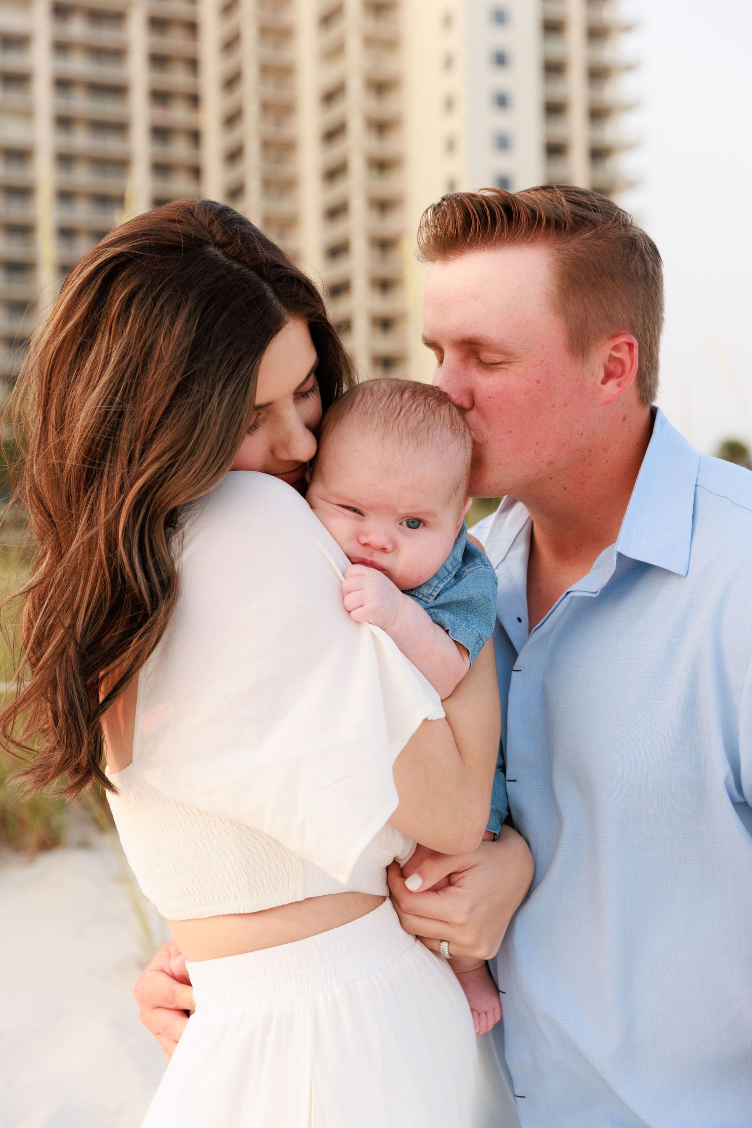 Young couple kissing newborn baby on beach with ocean resort building in background.