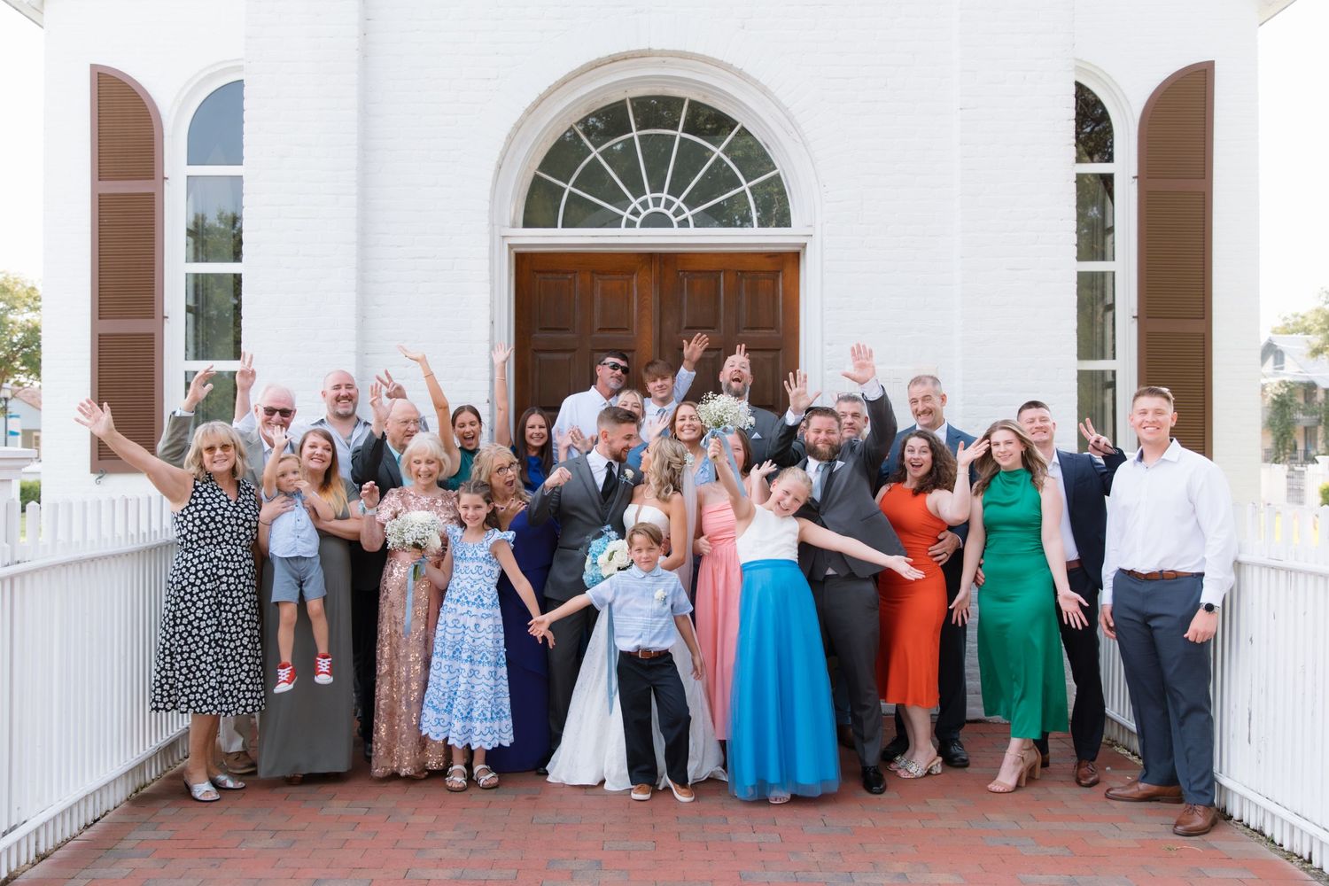 Large wedding party gathered outside white church building with arched doorway for group photo.