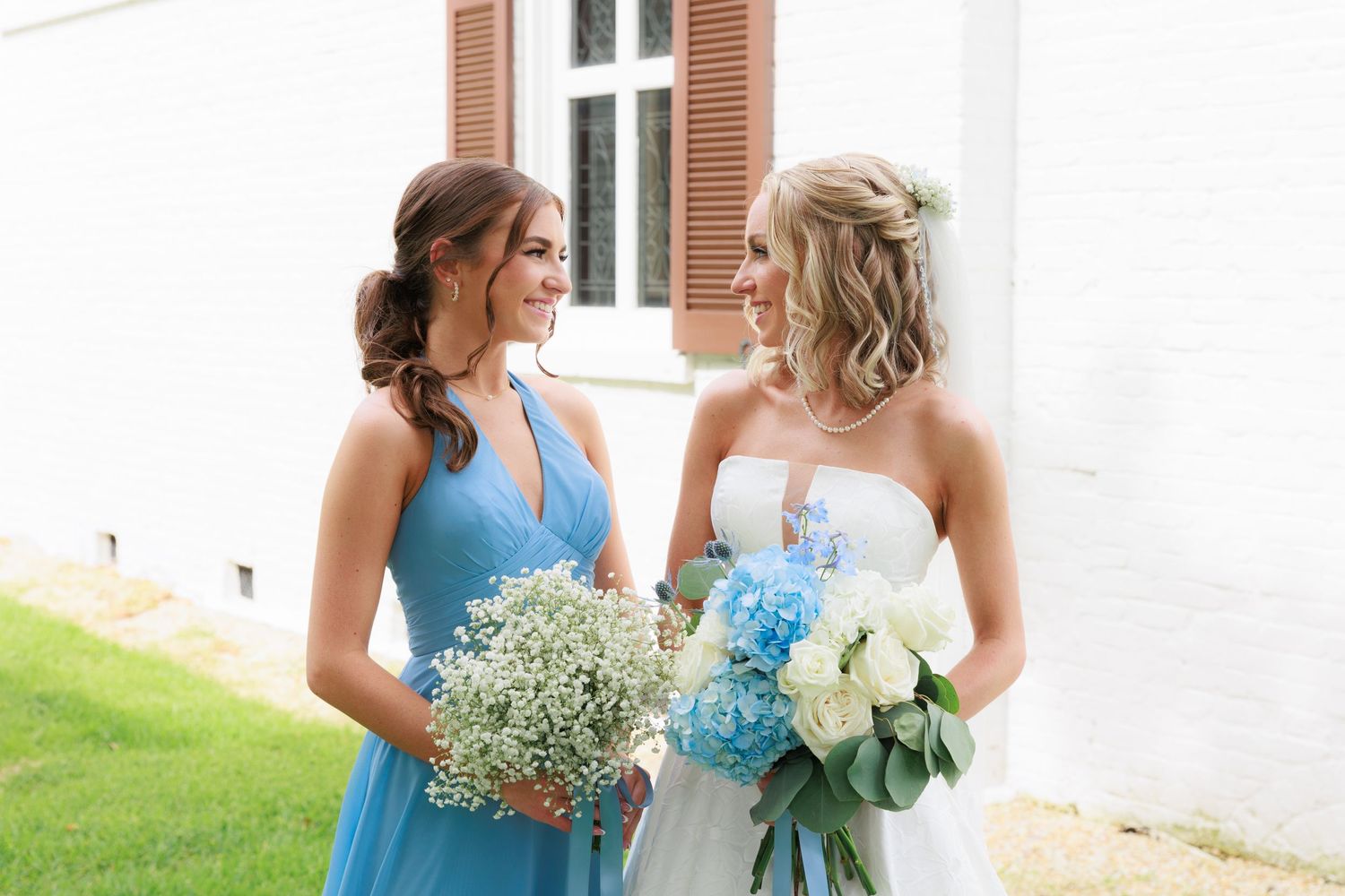 Bridesmaid in blue dress and bride share a moment while holding floral bouquets.
