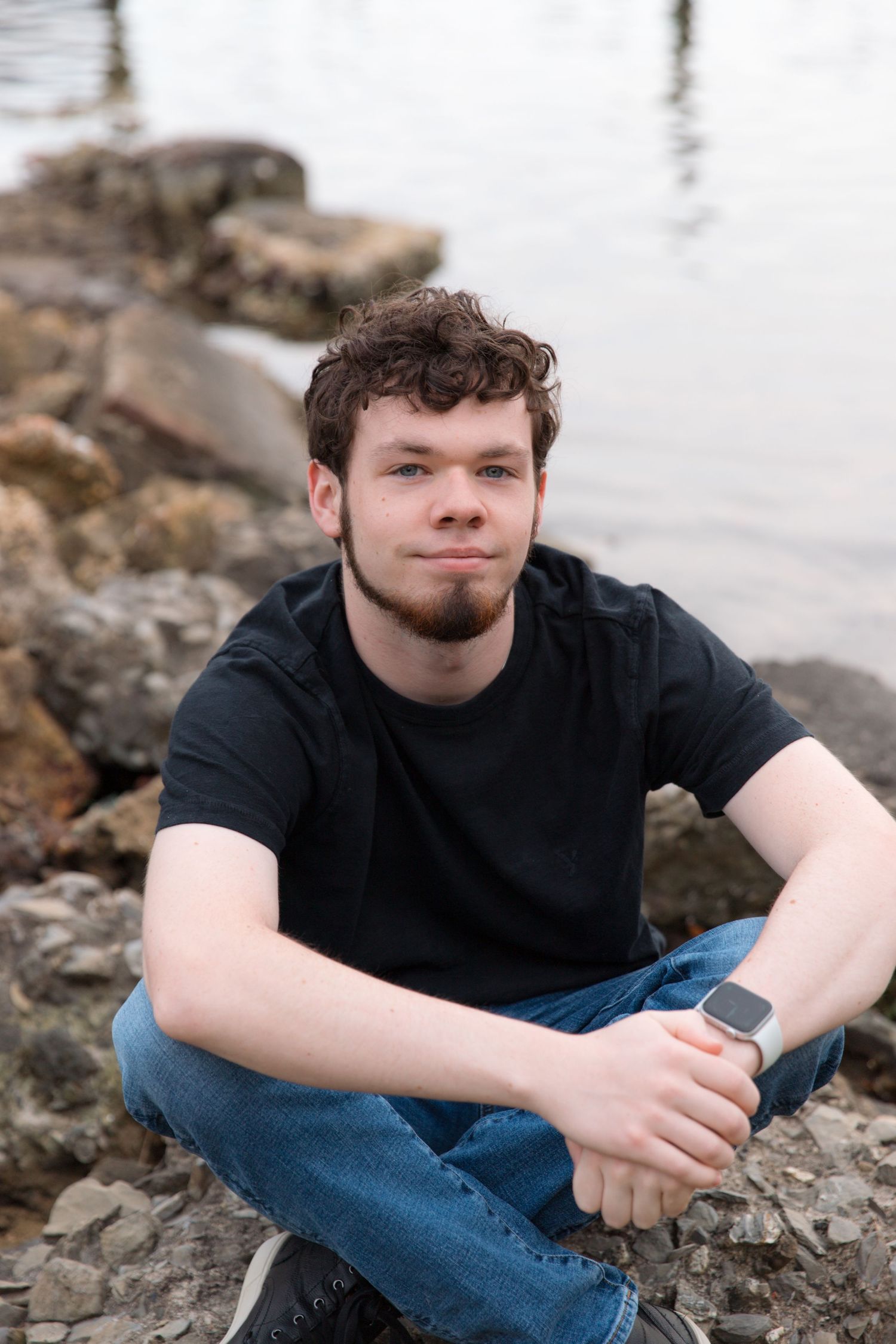 Sitting on rocky shoreline wearing black t-shirt and jeans in casual pose.