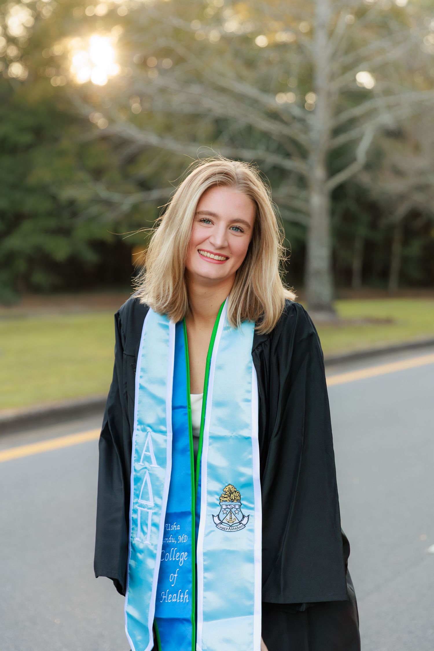 Graduate wearing black academic regalia and blue stole smiles during outdoor photo session.