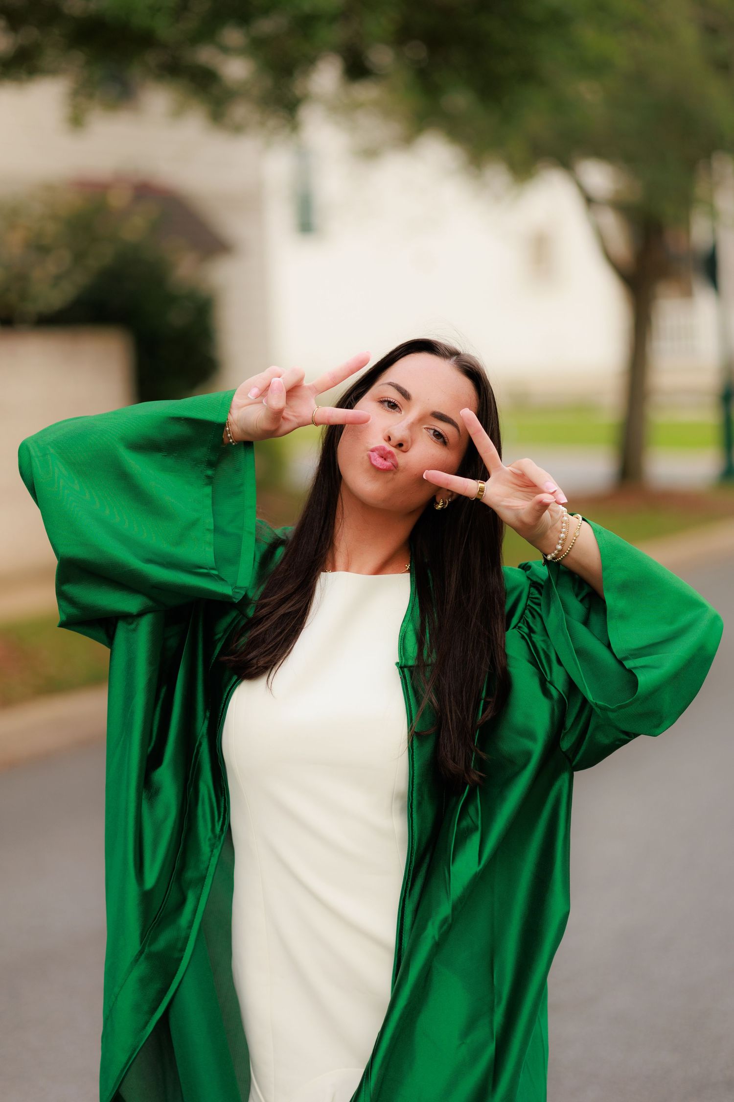 Young woman in green graduation gown making peace signs, wearing white dress, celebrating outdoors.