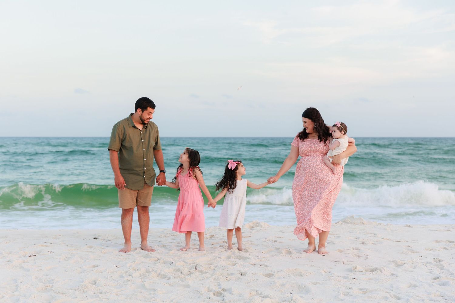 Family of five holding hands and walking on a sandy beach with turquoise ocean waves at sunset.