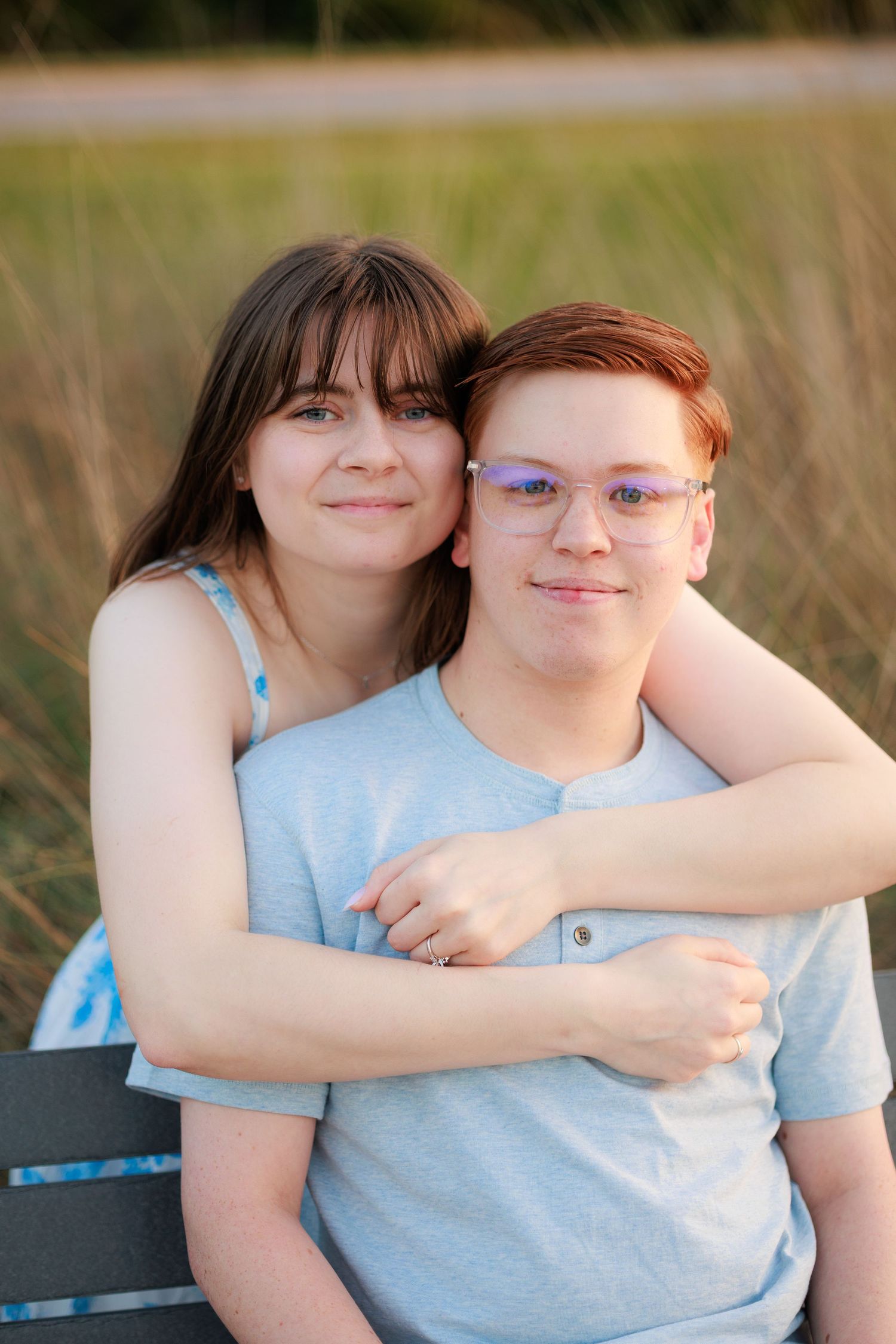 A young couple sits on a bench outdoors, the girl hugging the boy from behind, both smiling at the camera.