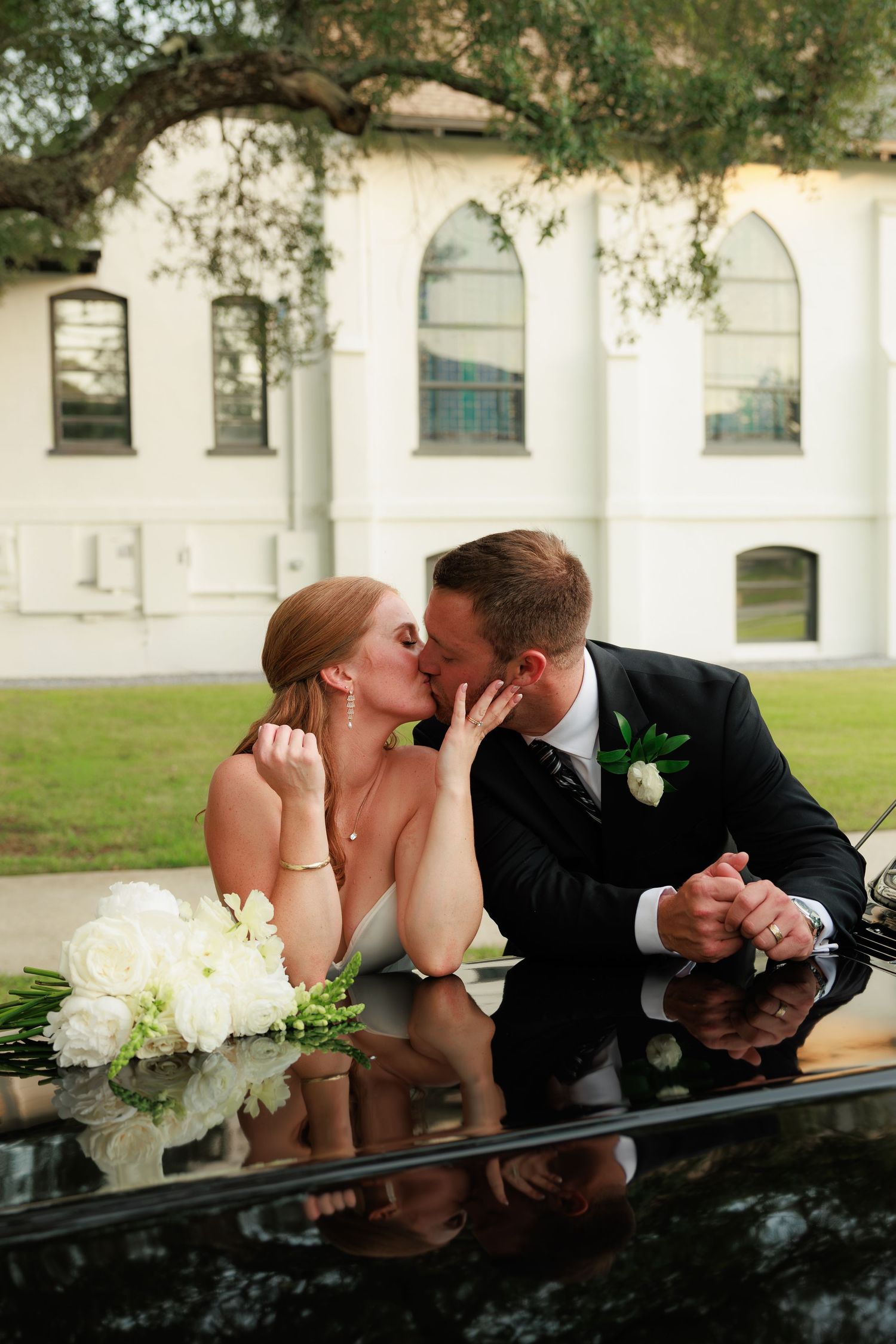 Bride and groom sharing a kiss leaning on black car hood with white bouquet outside white church venue.