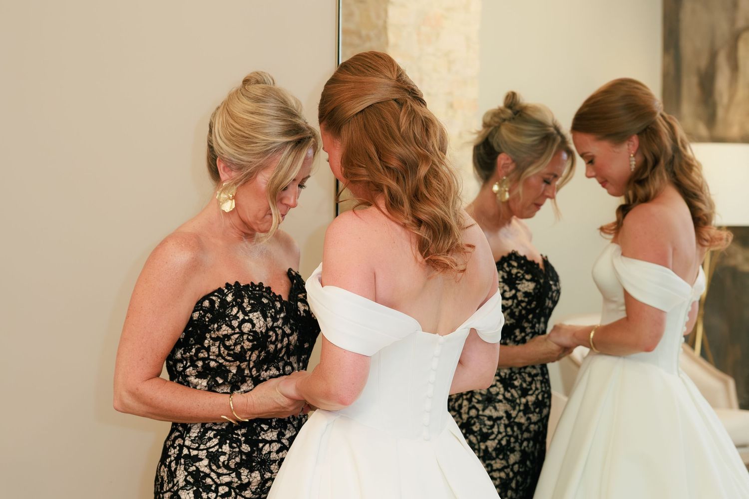 Bride in white off-shoulder wedding gown being helped by two women in floral dresses during bridal preparation.