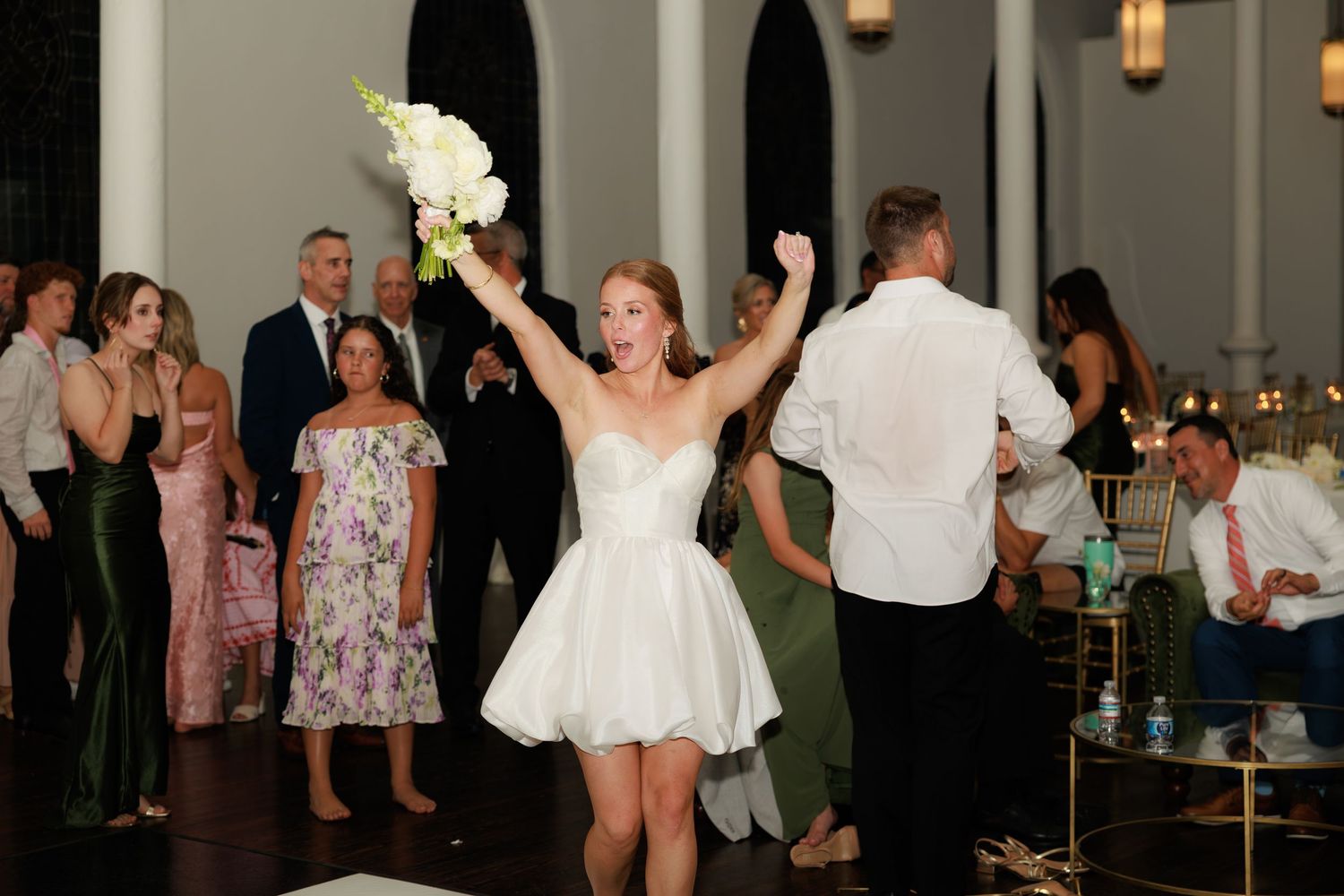 Bride in white mini dress raises bouquet triumphantly at wedding reception surrounded by guests.