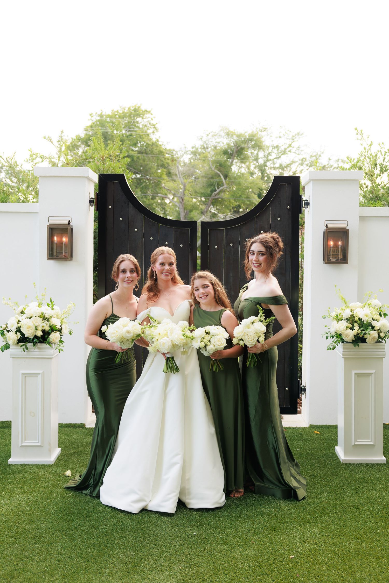 Bride in white gown with three bridesmaids in emerald green dresses holding white bouquets before elegant black gates.