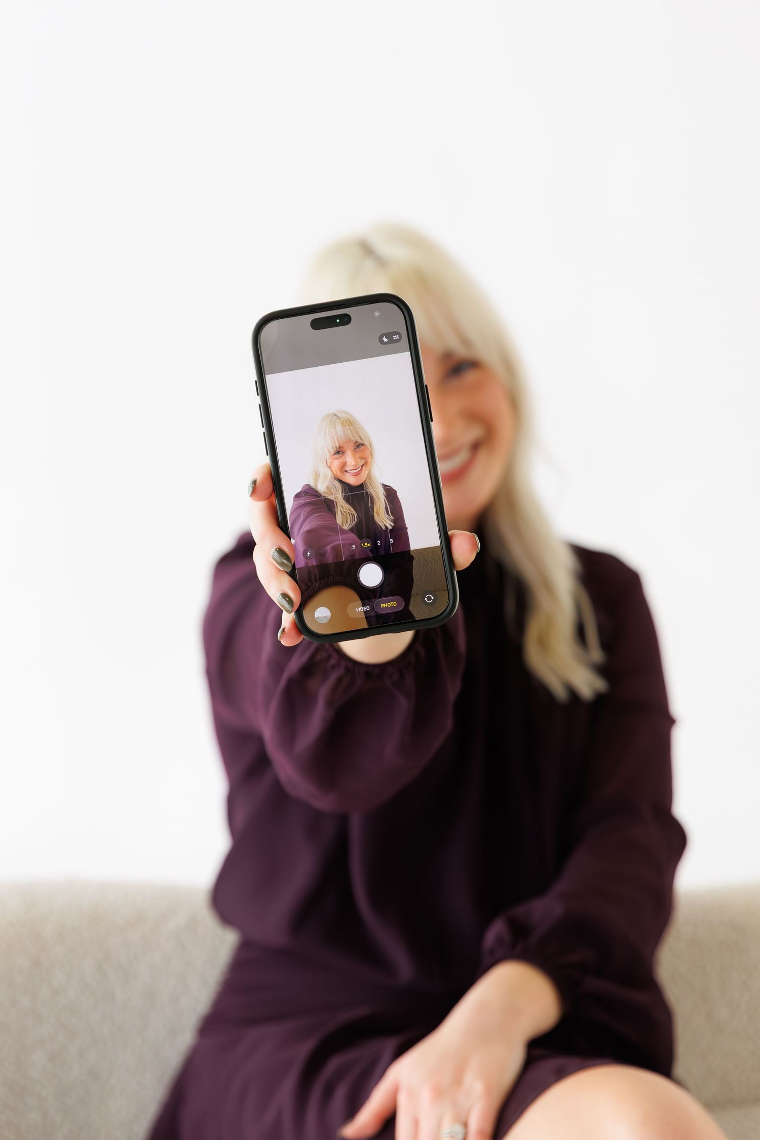 Person taking selfie with phone while sitting on couch in purple sweater dress.
