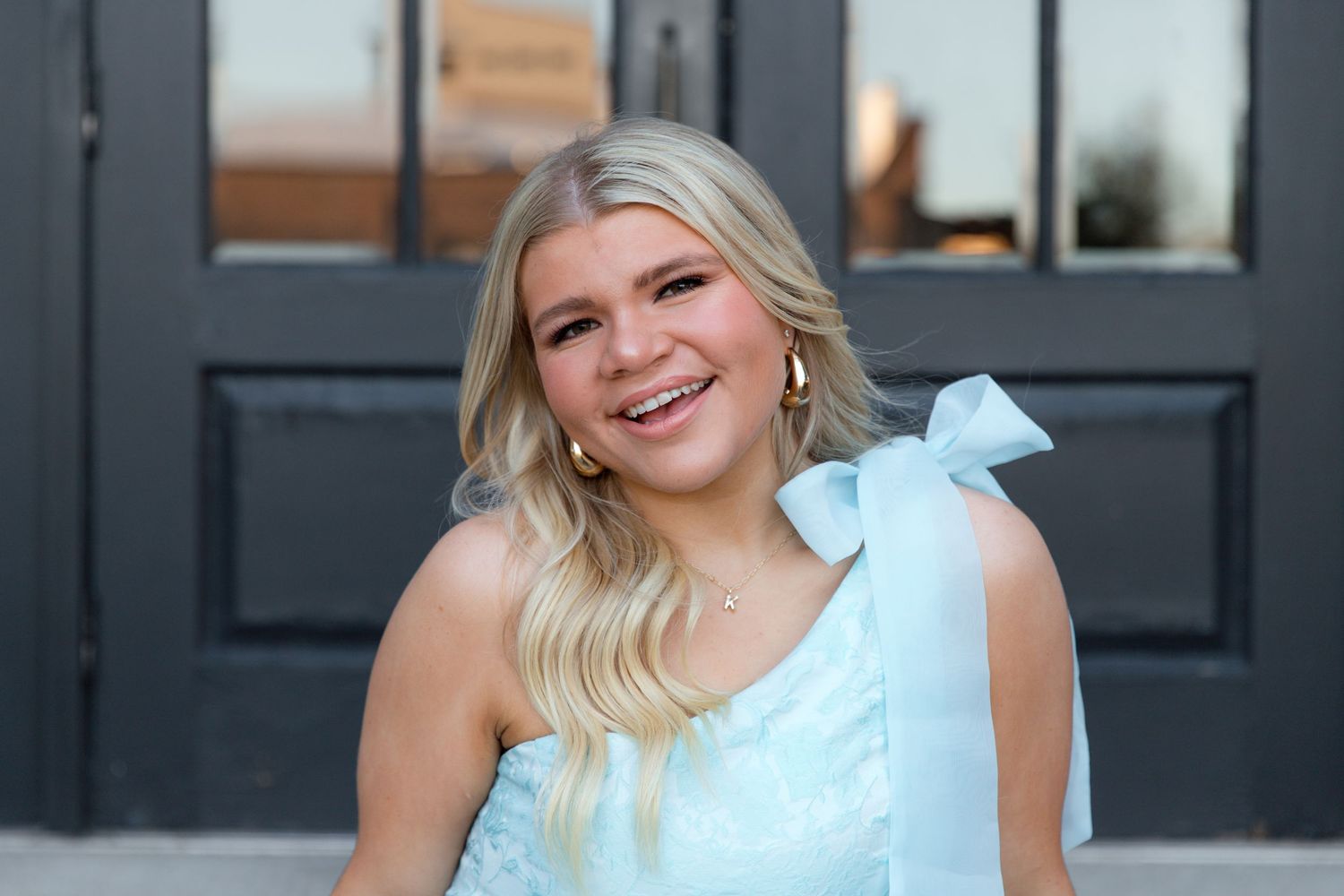 A person wearing a light blue dress with bow detail poses in front of a dark door.