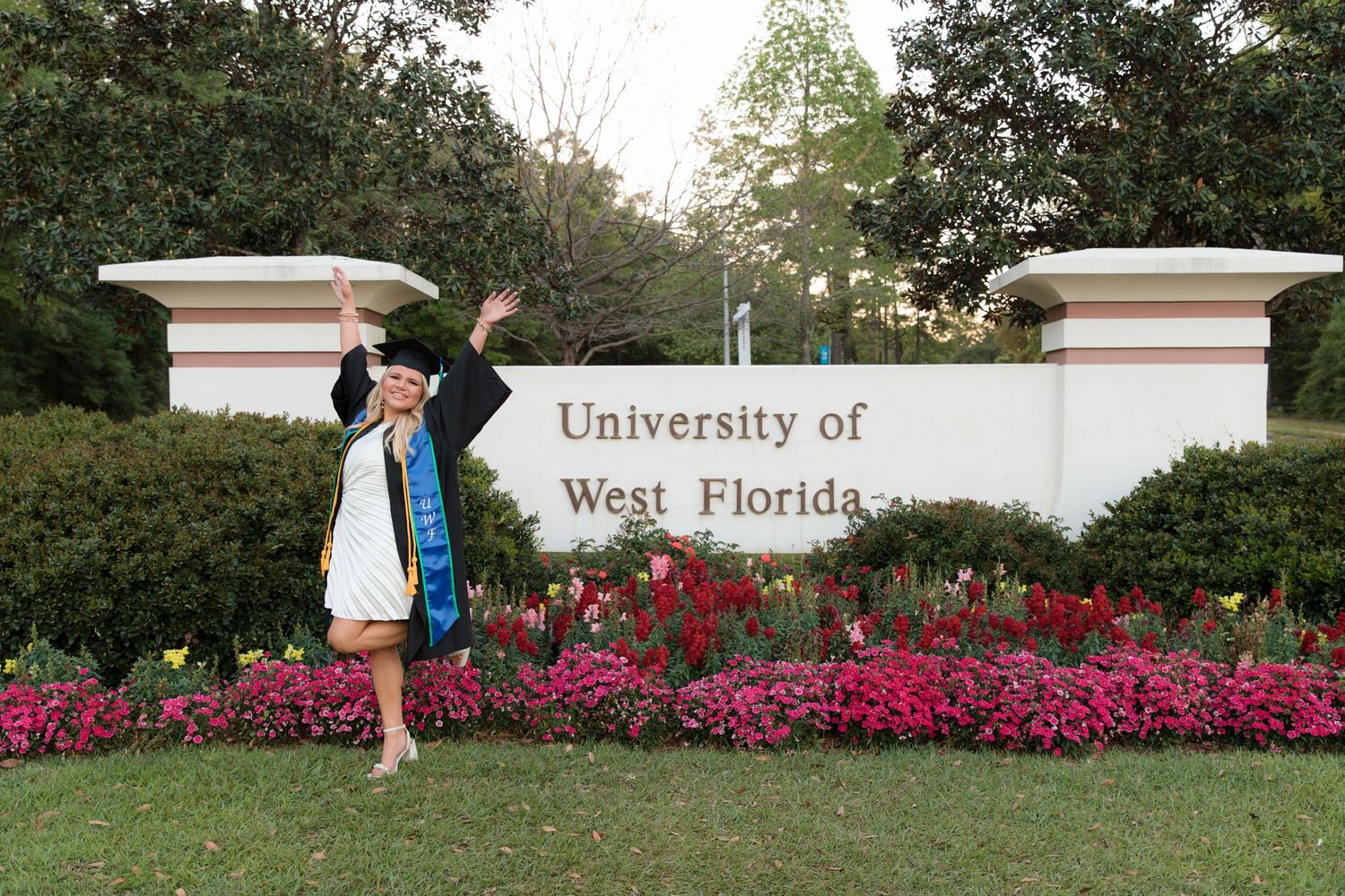 A graduate poses next to the University of West Florida entrance sign surrounded by bright pink flowers.