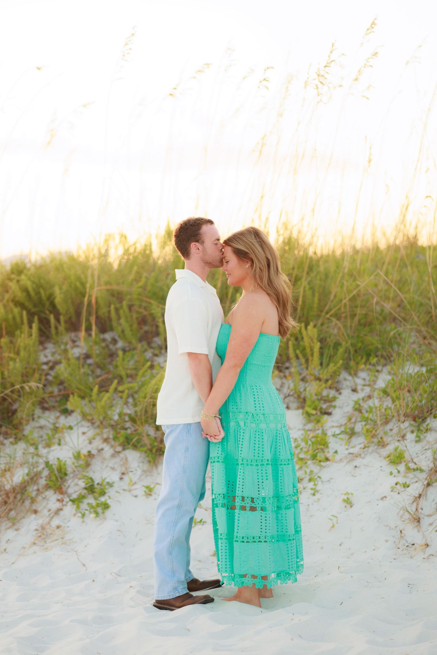 A couple shares tender moments on a sandy beach with sea oats swaying in the golden evening light.