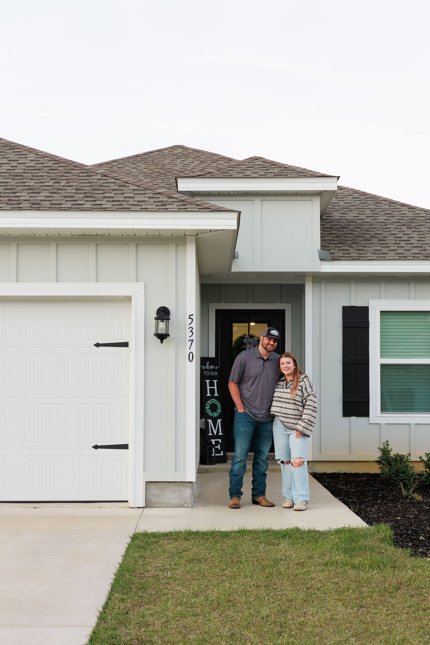 Two people stand in the doorway of a white residential house with green shutters and a concrete driveway.