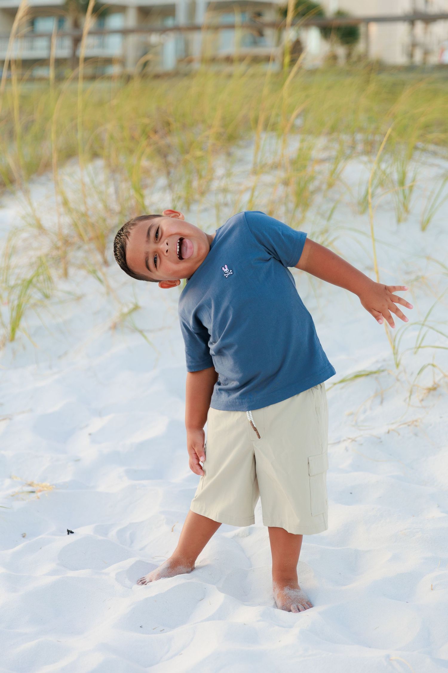 Child in blue shirt and khaki shorts plays joyfully in pristine white sand on a sunny beach.
