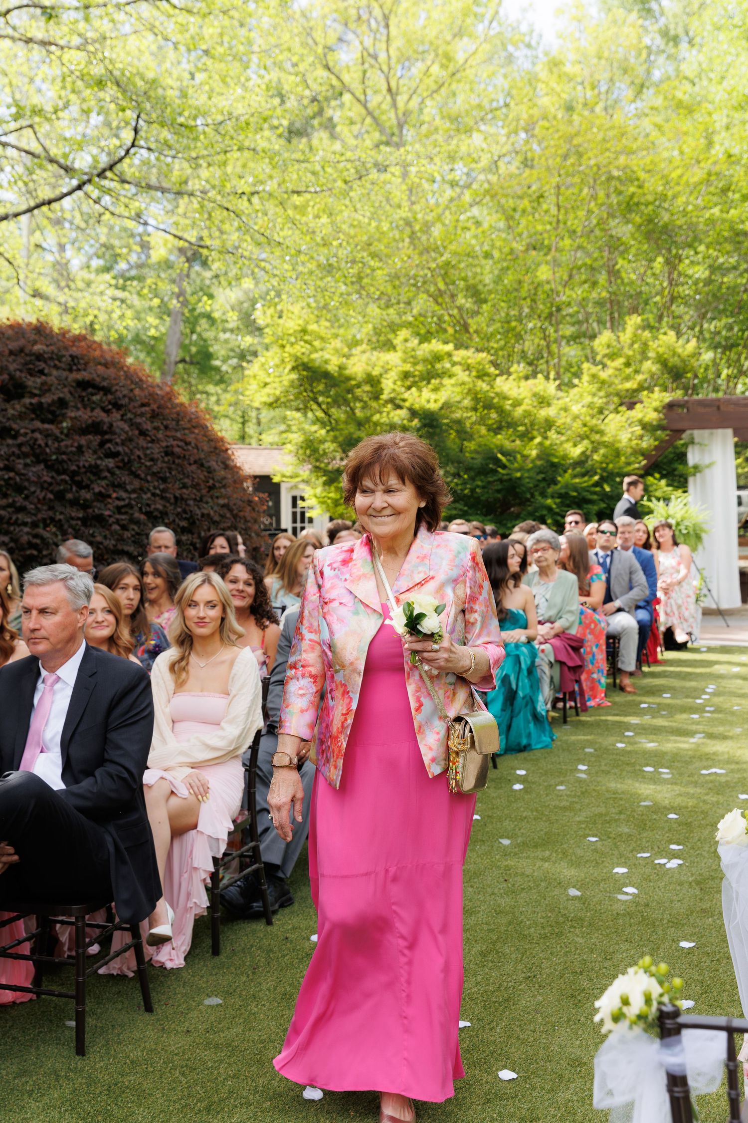 Woman in pink dress and floral jacket walks down outdoor wedding aisle holding white flowers.