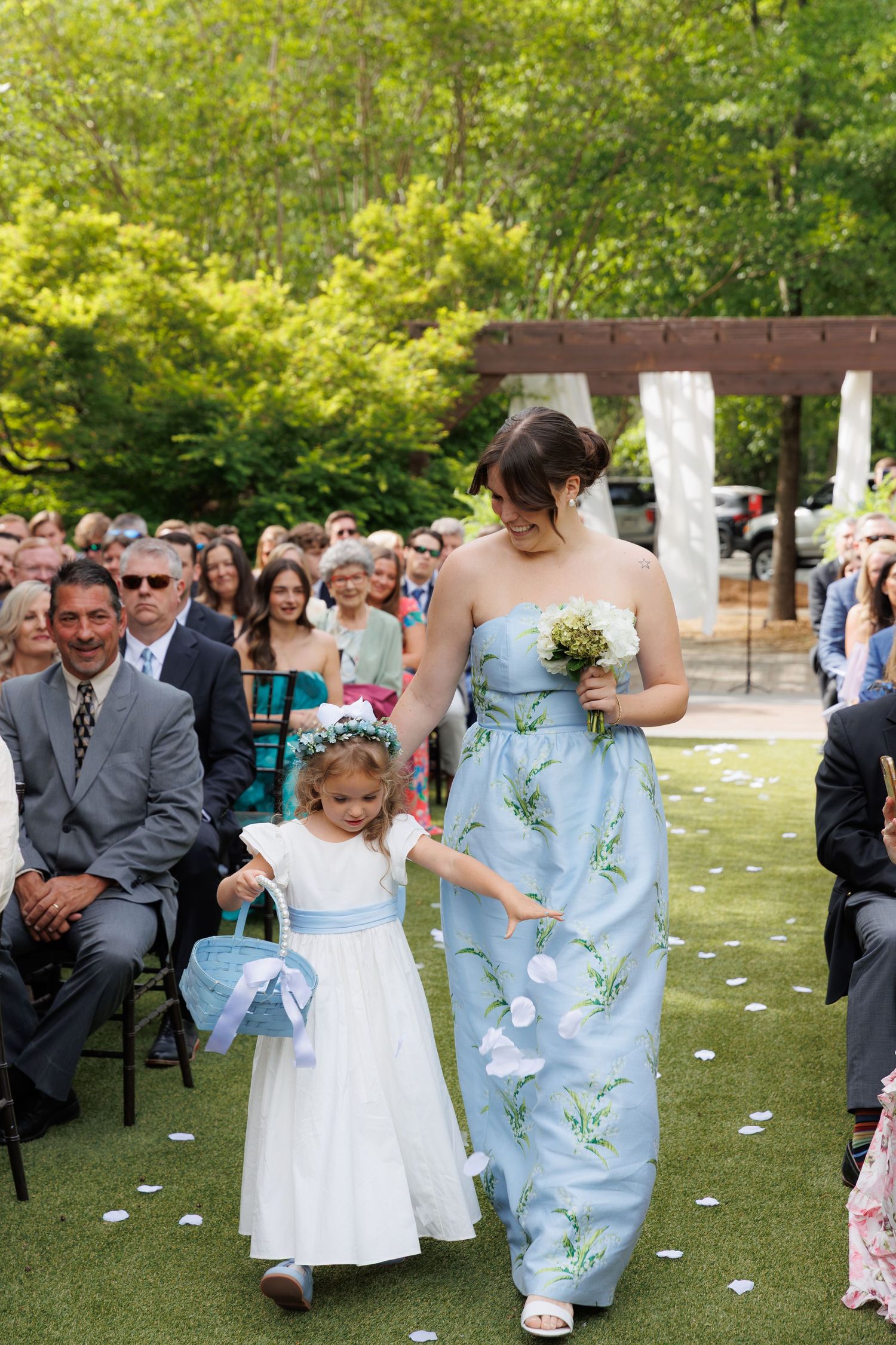 Bridesmaid in floral blue gown walks flower girl in white dress down outdoor wedding aisle amid seated guests.